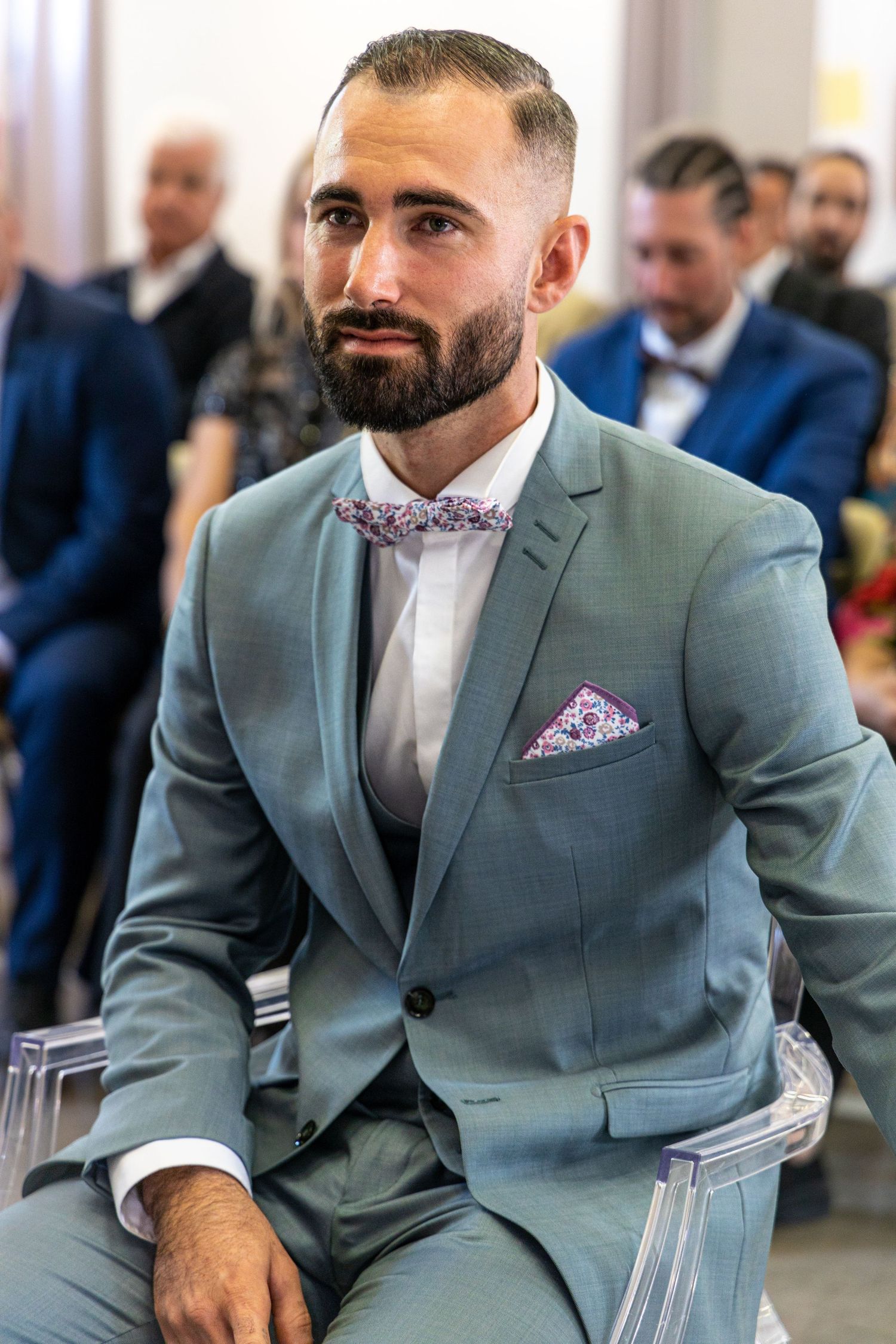A groom in a stylish mint green suit with floral bowtie sits during wedding ceremony.
