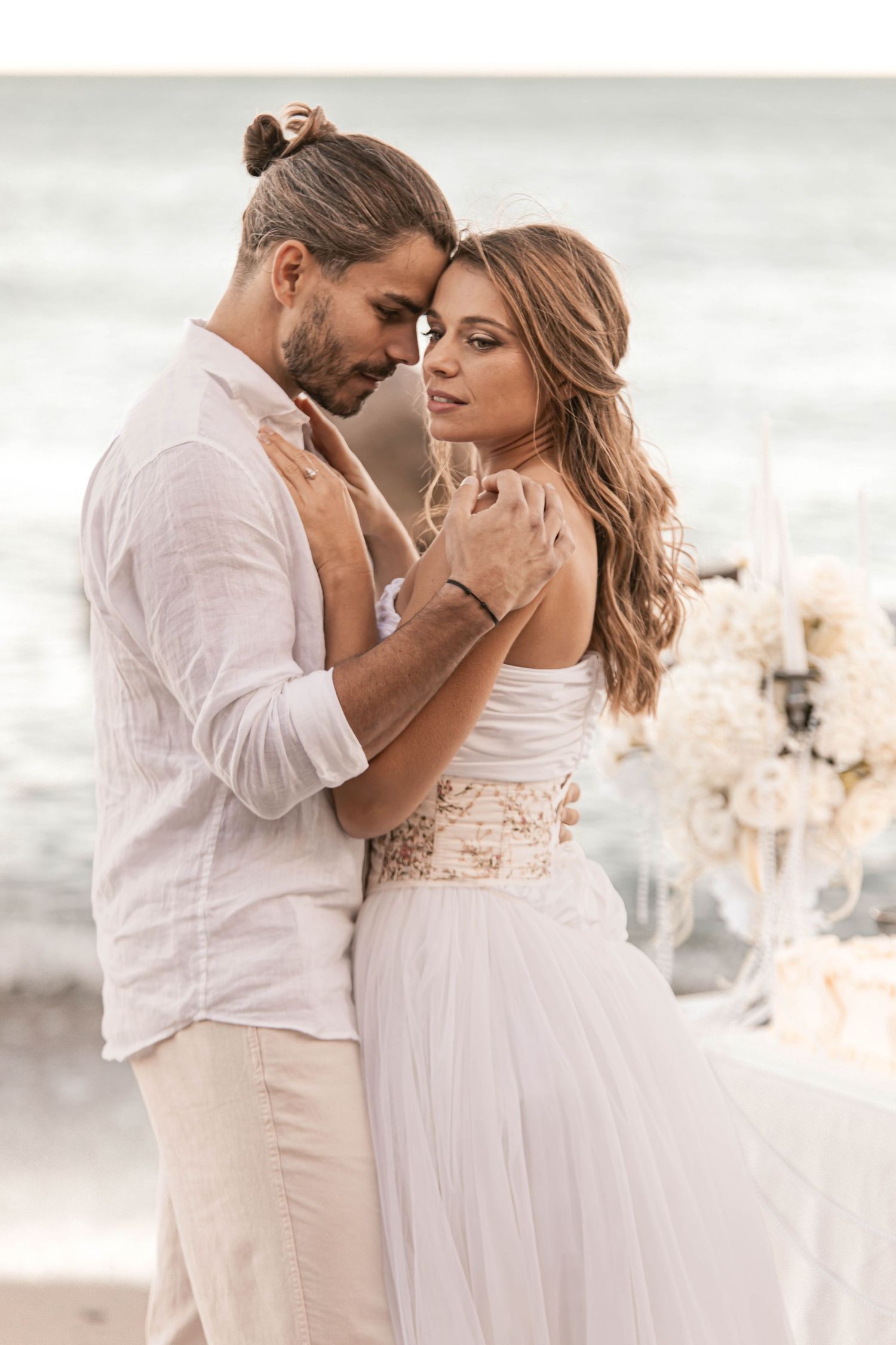 Couple in white attire embracing tenderly on a beach, with floral arrangements in the background at a seaside wedding.