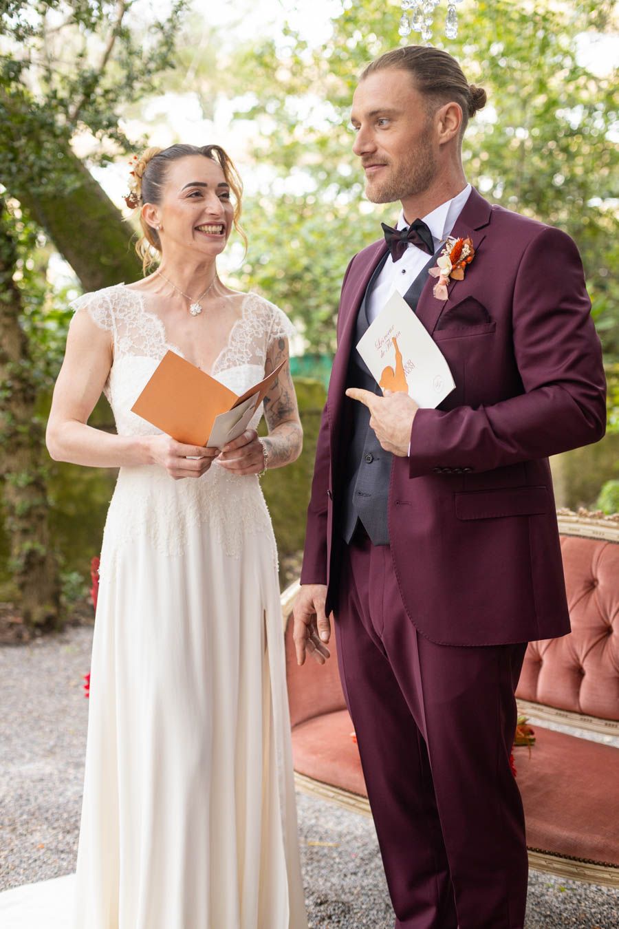 Wedding couple in burgundy suit and white dress share joyful moment holding orange cards.