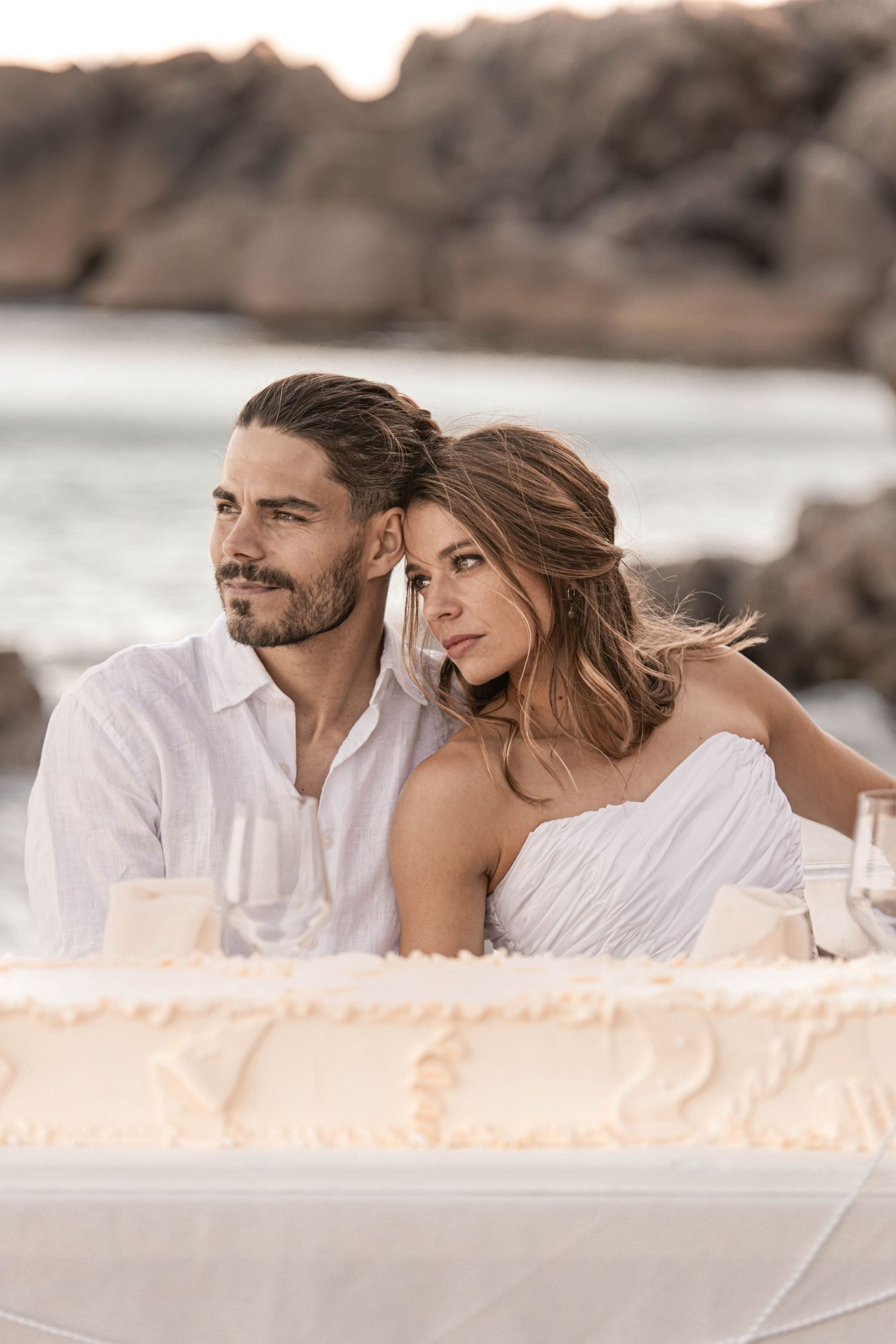 Romantic couple in white attire sitting together at a beachside table with a wedding cake at sunset.