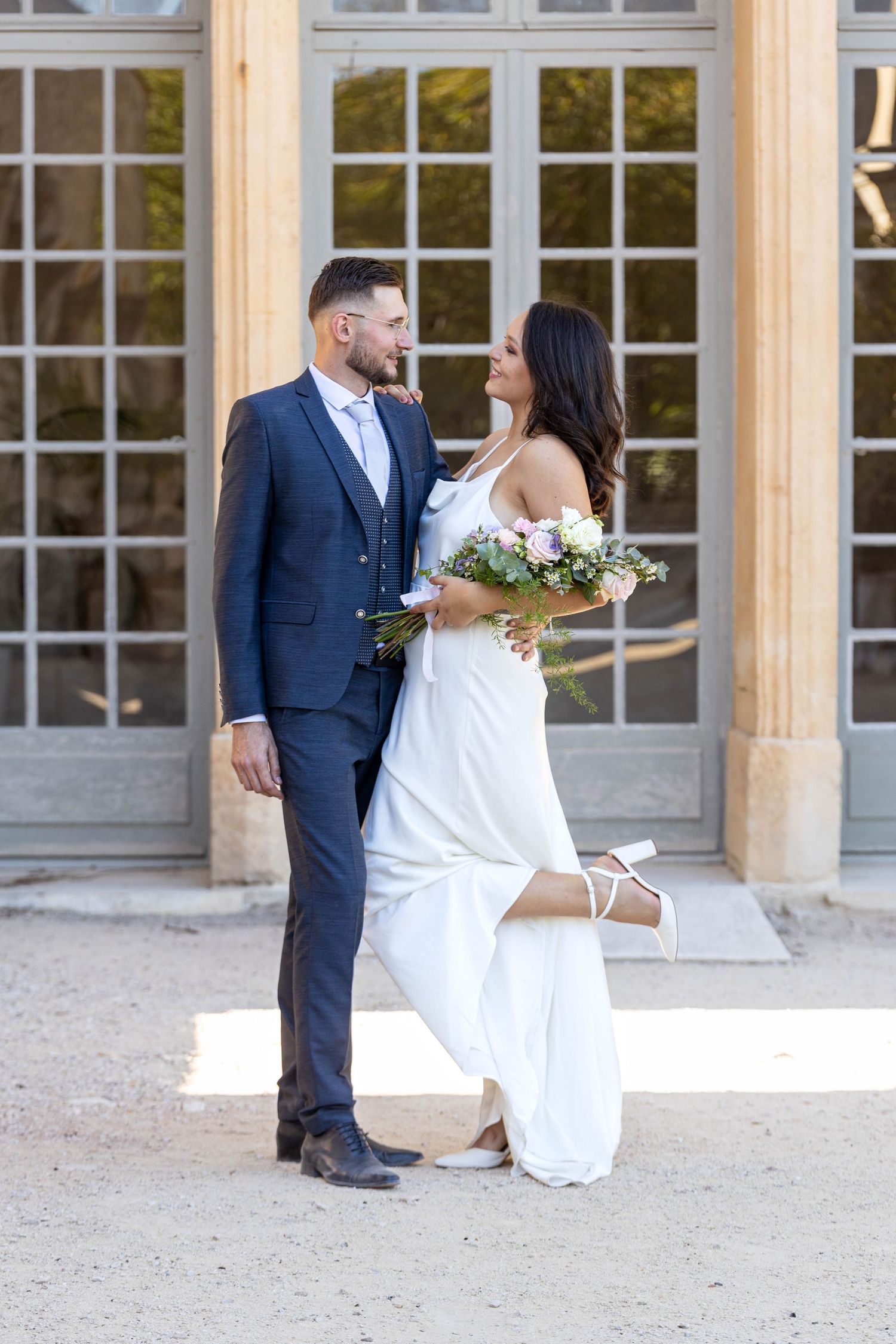 Bride in white gown and groom in navy suit share intimate moment by columned building.