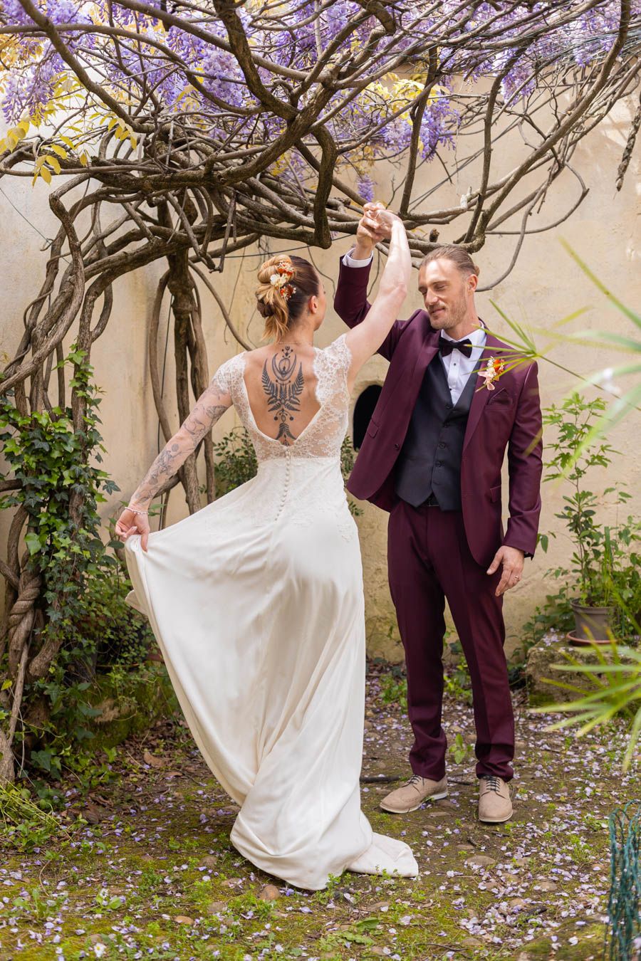 Wedding portraits under wisteria showing bride's lace dress details and groom's burgundy suit.
