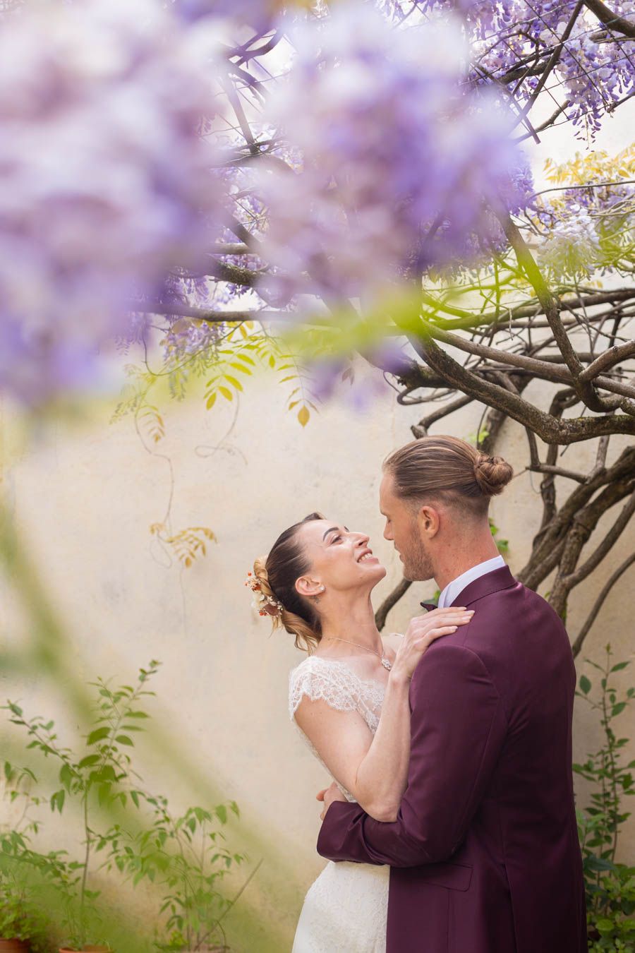 Wedding portrait framed by delicate purple wisteria blossoms in a garden setting.