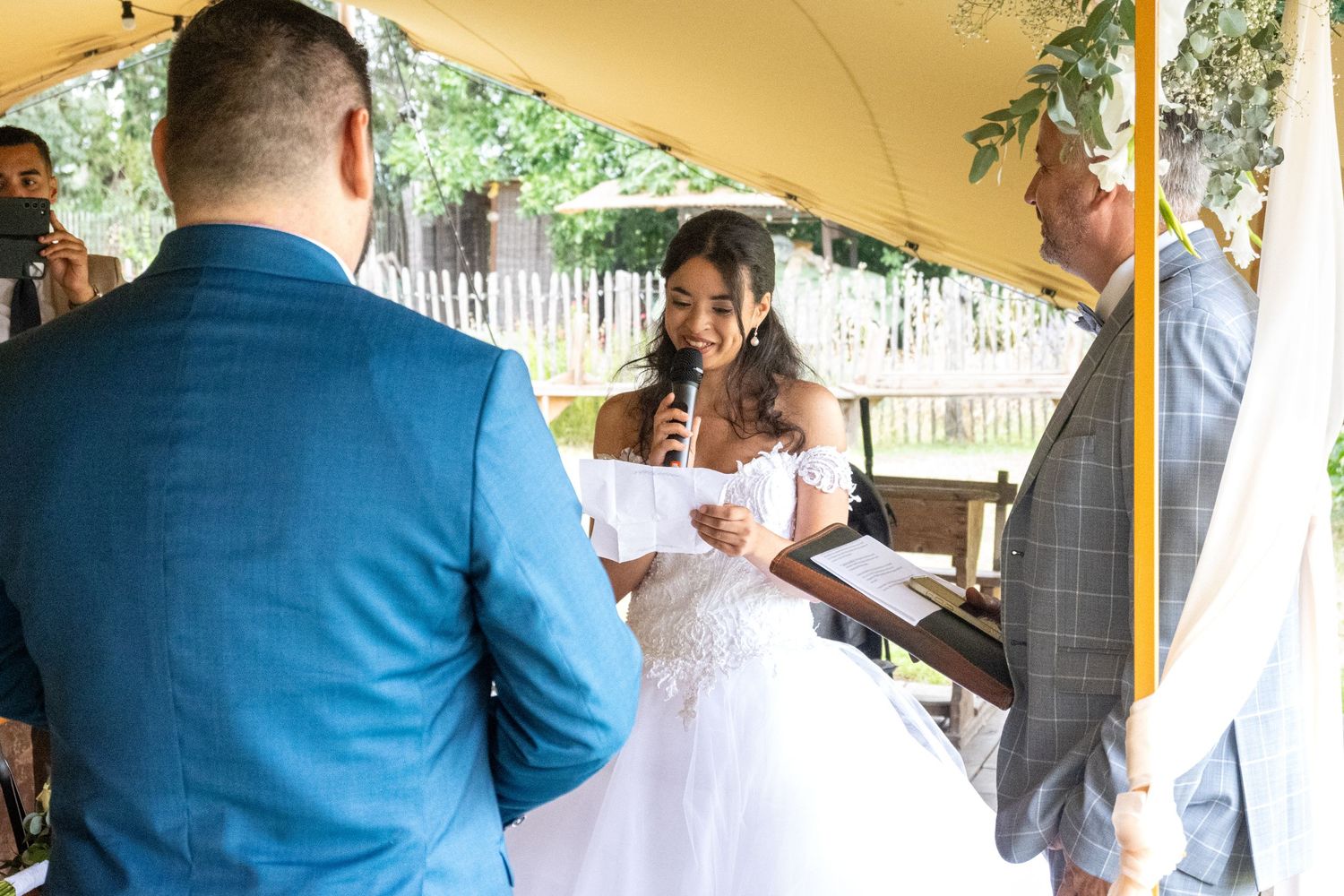 Outdoor wedding ceremony under a beige tent with guests seated in white chairs.
