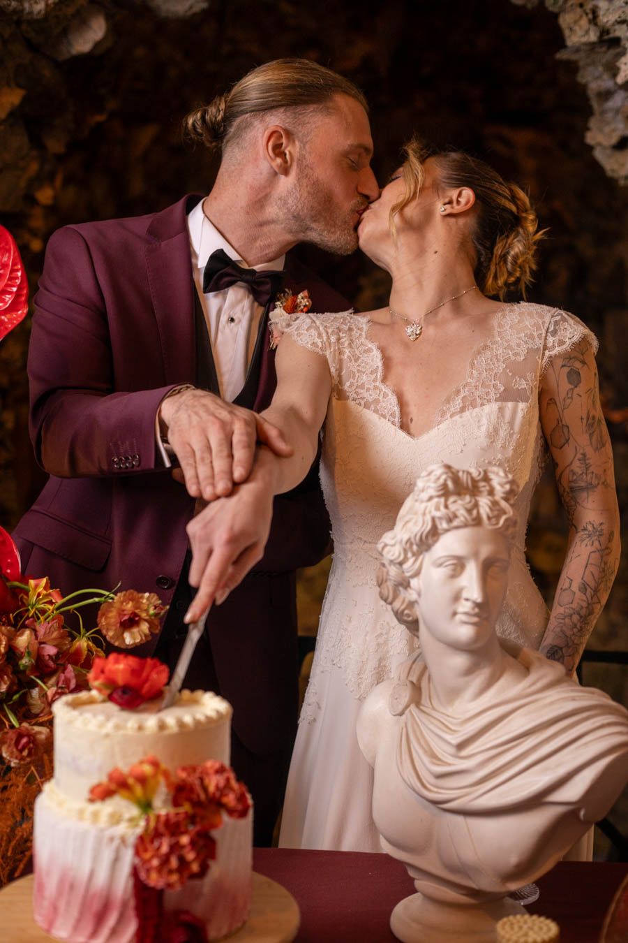 Wedding cake cutting scene with classical bust sculpture and red floral decorations.