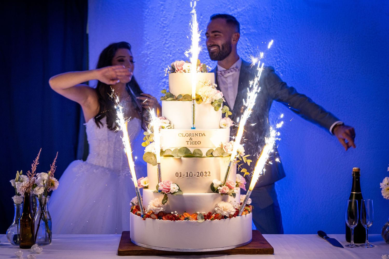 Wedding cake with sparkler fountains against vibrant blue backdrop during celebration.