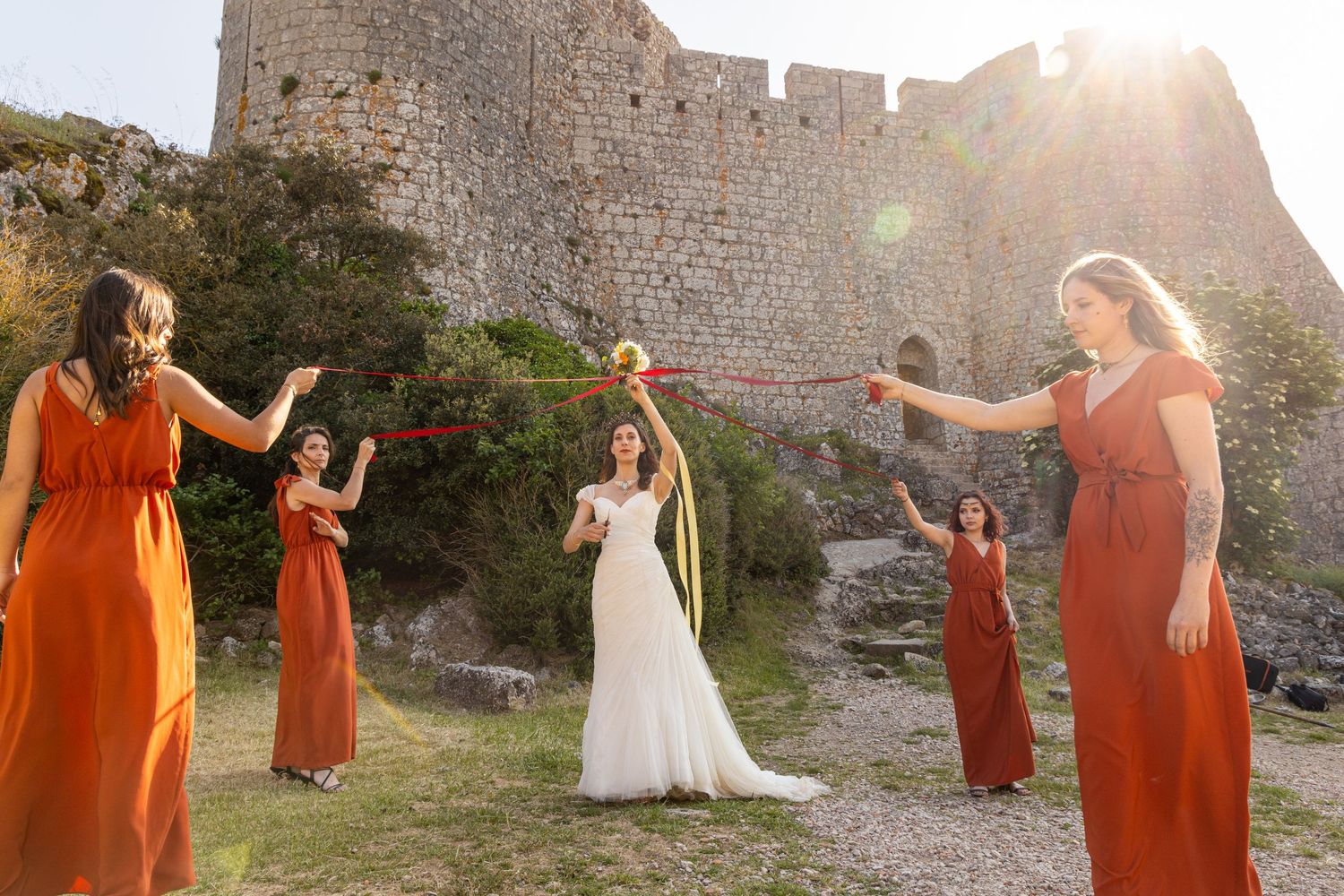 Creative wedding photo with bridesmaids in orange dresses near a castle wall at sunset.