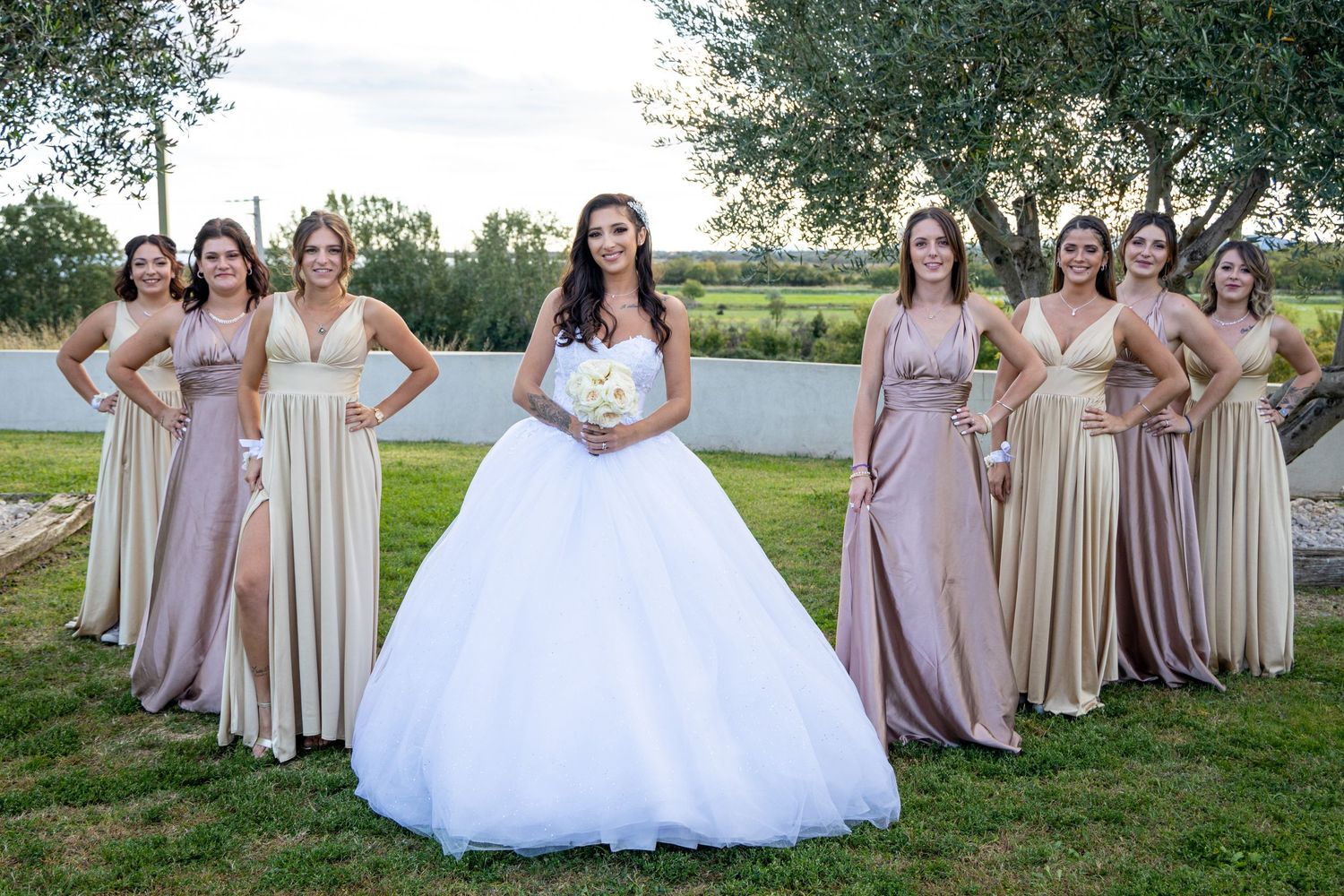 Bridesmaids in neutral-toned dresses pose with bride in white ballgown in an outdoor garden setting.