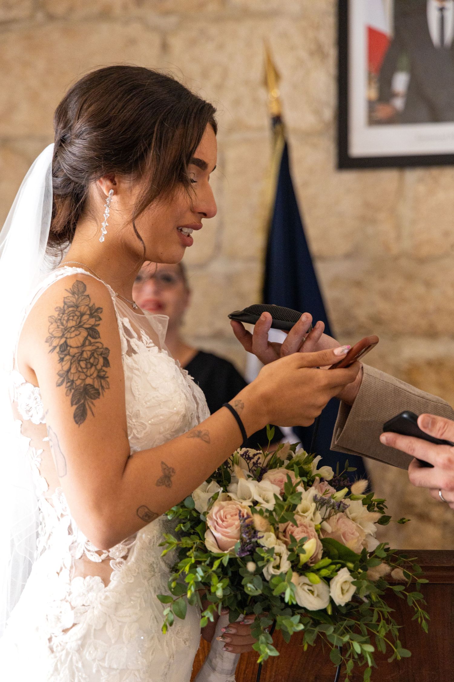 Bride in lace dress with floral tattoo smiles while holding something during wedding ceremony.