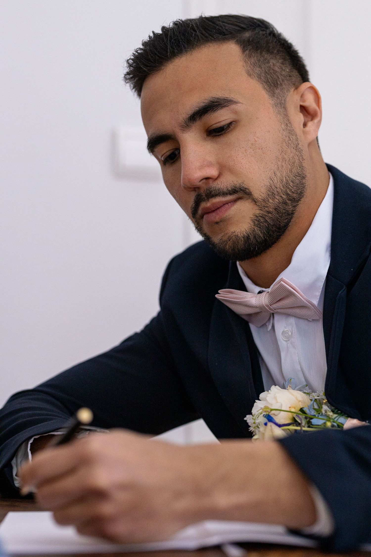 Someone in a navy suit and white shirt signs documents with a boutonniere on their lapel.