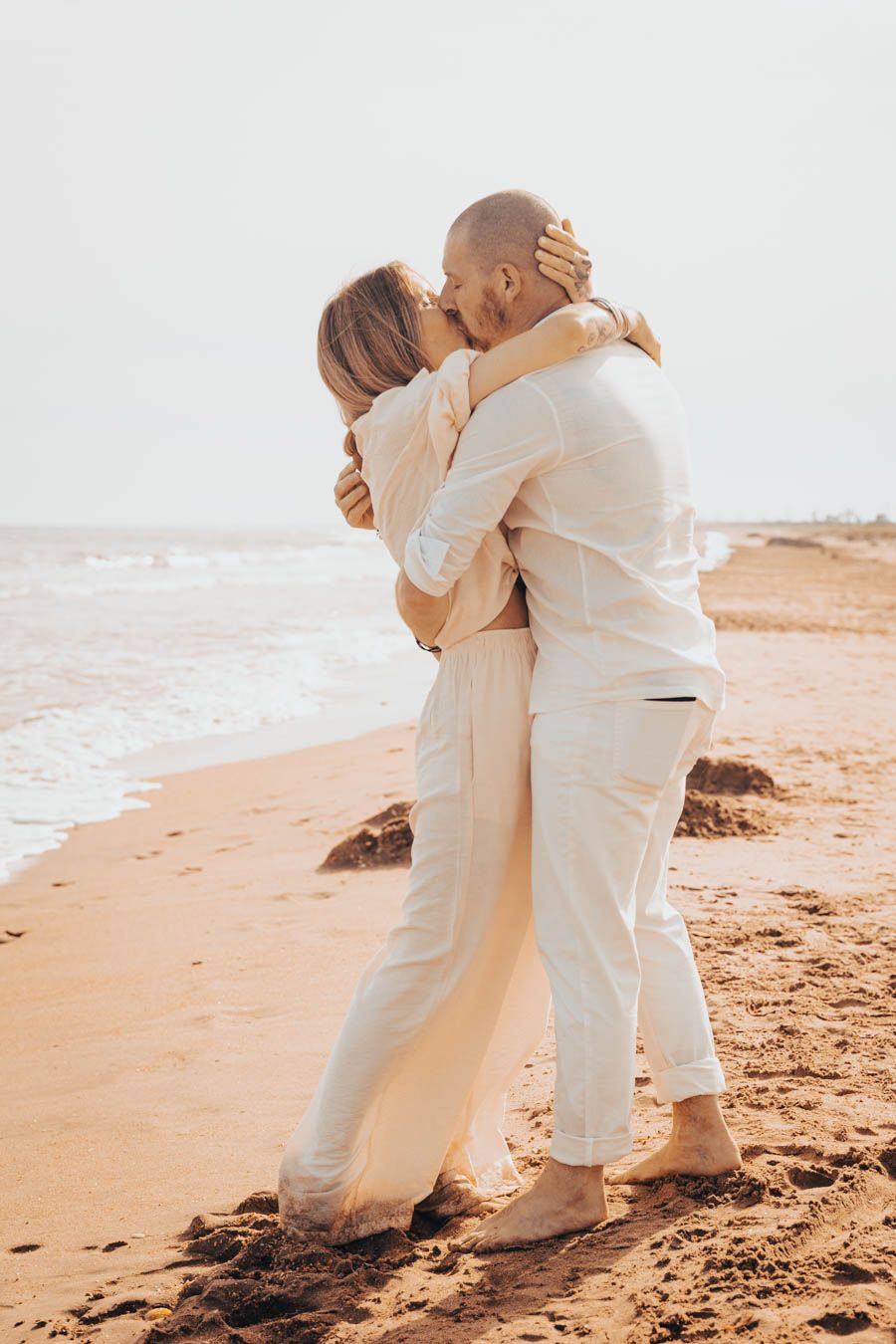 A couple in white outfits embraces on a sandy beach at sunset in a romantic beach photoshoot.
