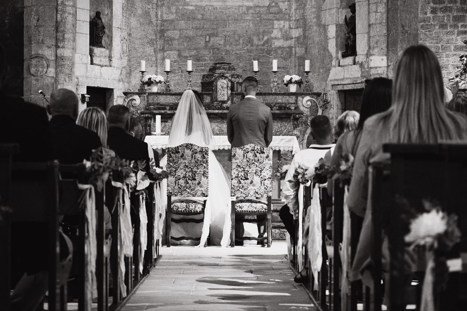 Wedding guests seated in wooden pews within a historic church with stone walls.