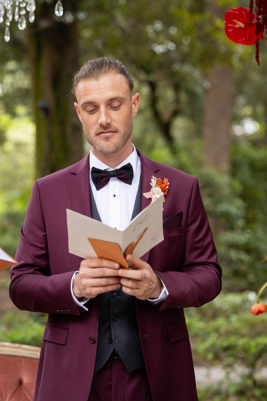 Person in burgundy tuxedo reads from card during outdoor wedding ceremony.