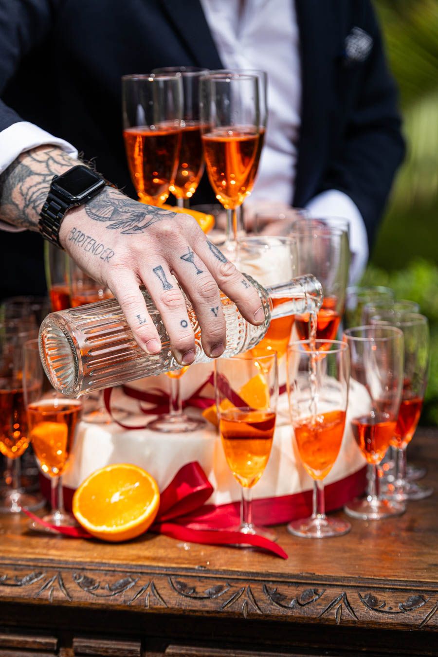 Tattooed hands prepare cocktails with orange garnishes on a rustic wooden bar surface.