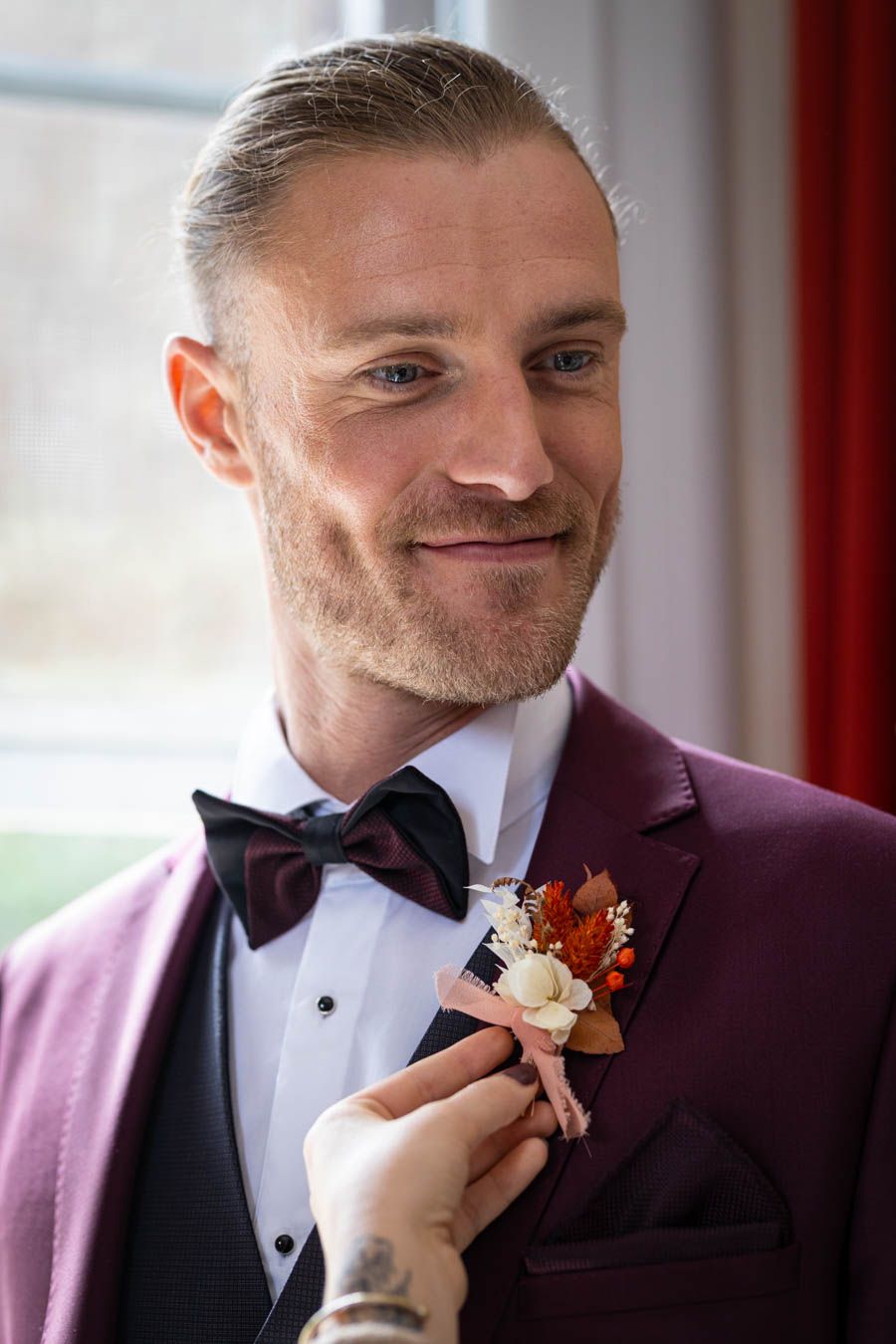 Burgundy tuxedo jacket with black bowtie and boutonniere being adjusted.
