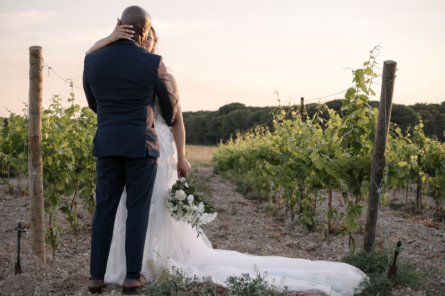 A couple walks along a scenic vineyard path at sunset in formal wedding attire.