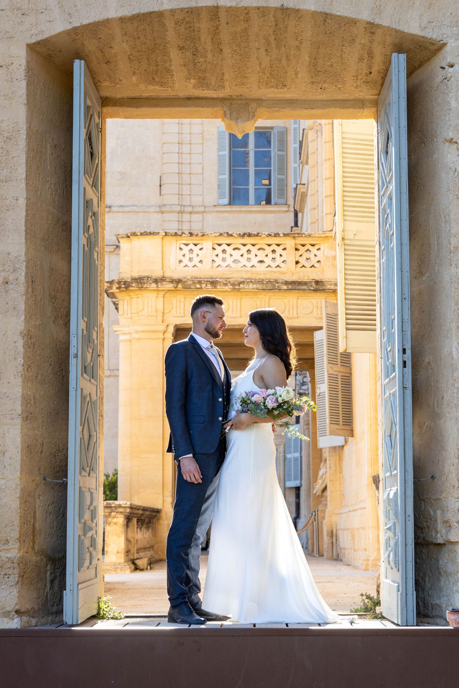 Wedding couple stands in an elegant colonnade with yellow shuttered windows.