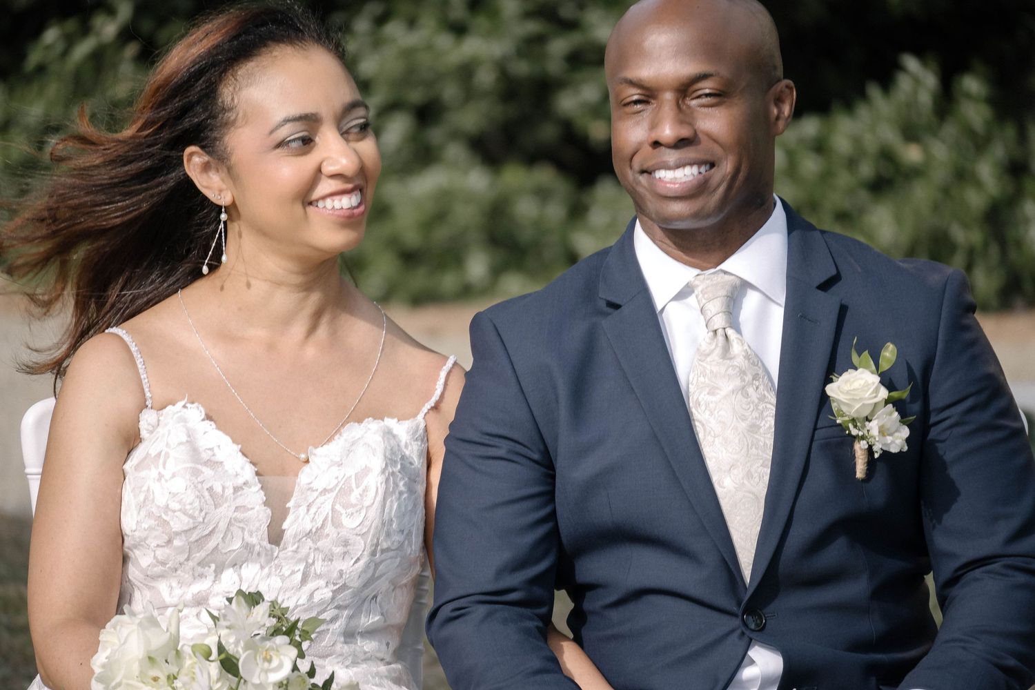 Wedding couple smiling and laughing together outdoors in formal attire with natural greenery backdrop.