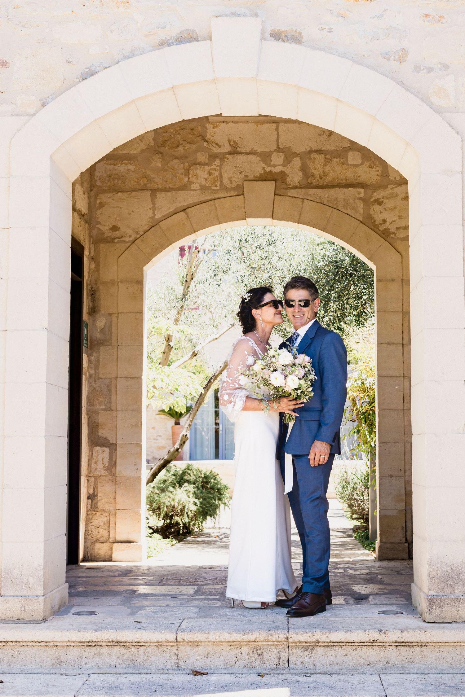 Wedding couple poses romantically under stone archways with elegant bouquet and formal attire.