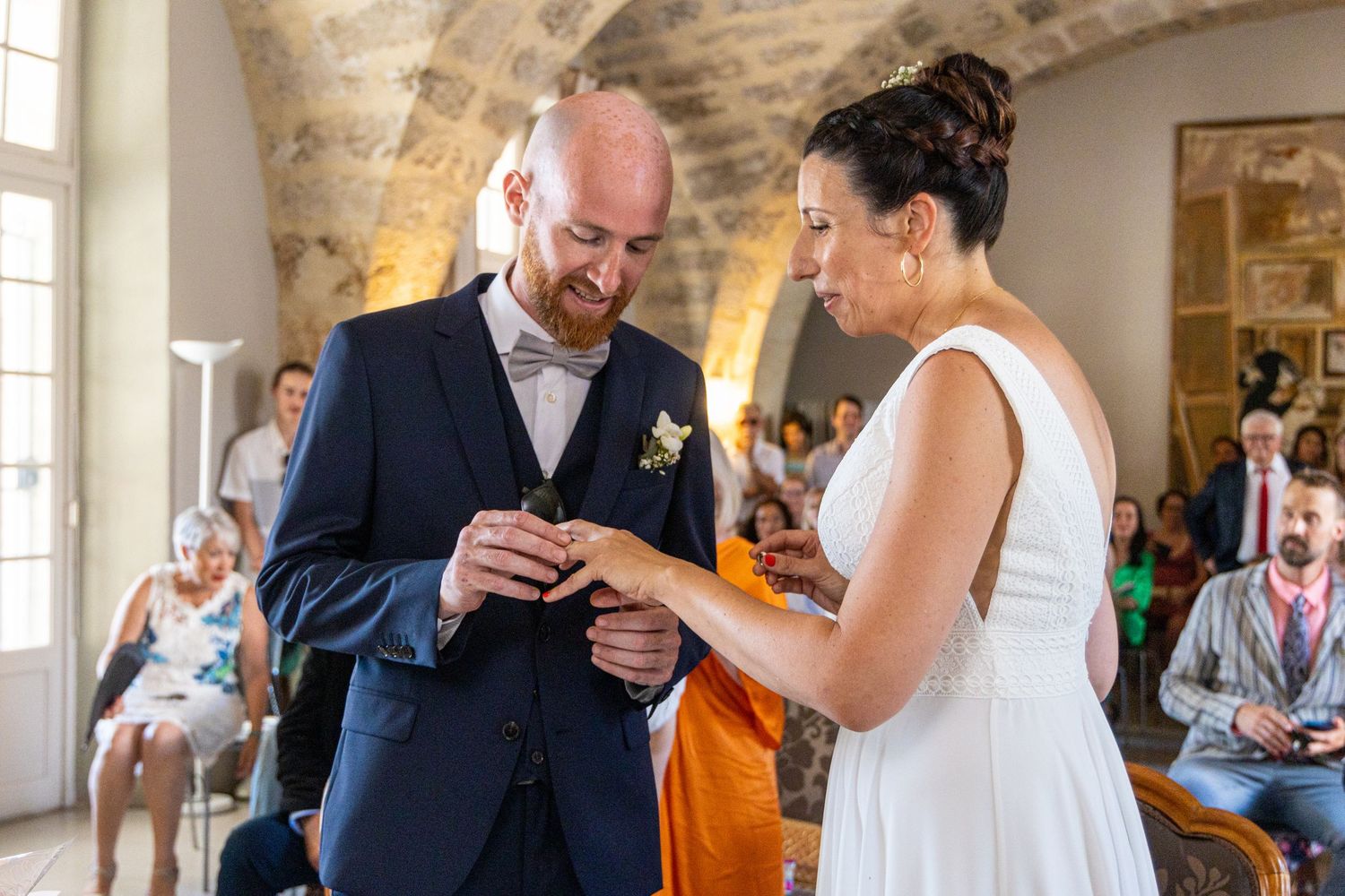 Wedding couple exchanges rings during intimate indoor ceremony with stone walls.