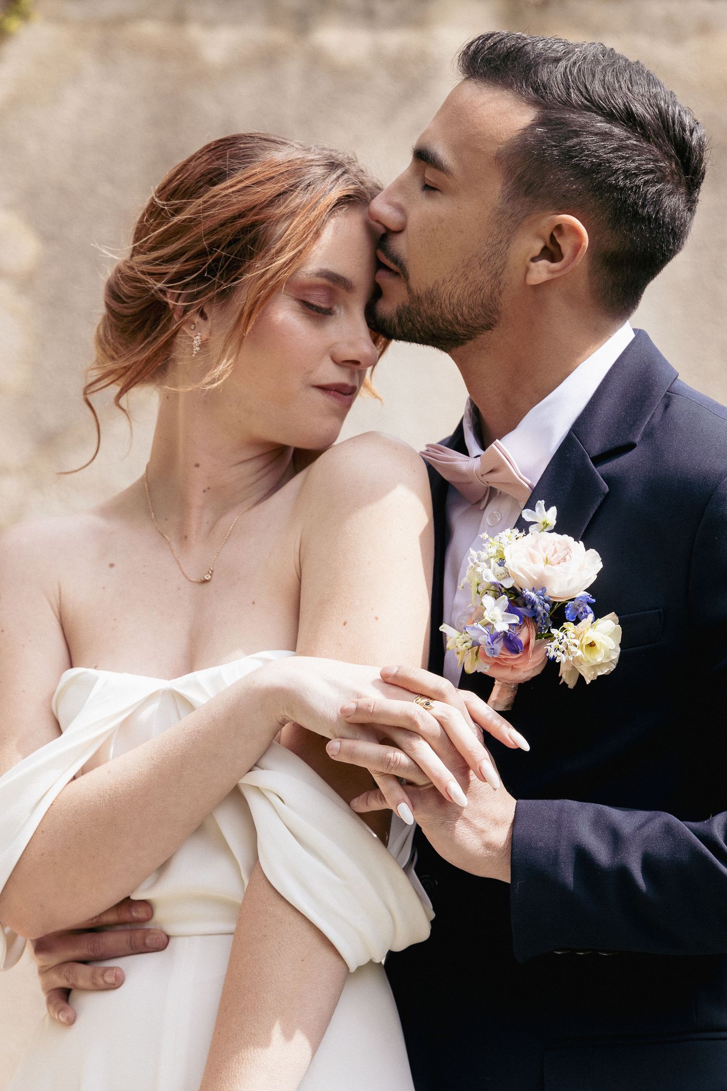 Intimate moment between newlyweds as groom kisses bride's temple with floral boutonniere.