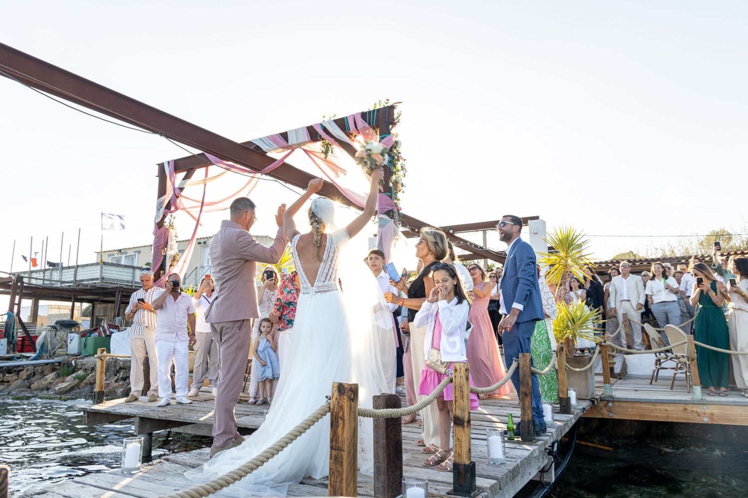 Wedding celebration on waterfront dock with floral arch and guests gathering at sunset.