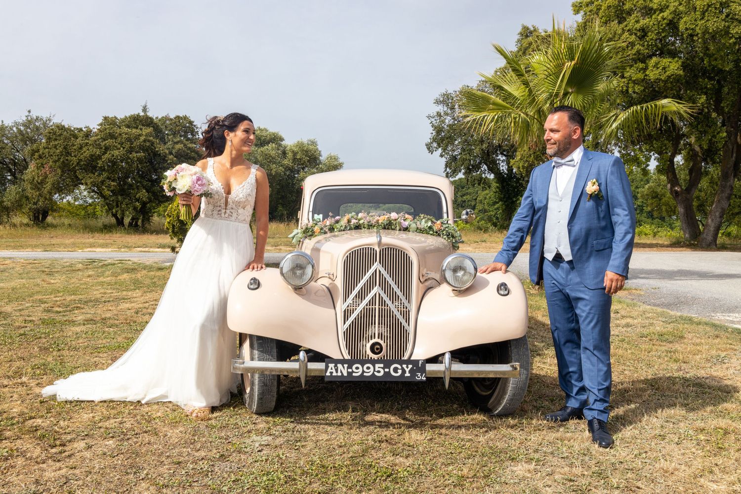 Newlyweds pose with vintage Citroen car in cream color against natural outdoor setting.