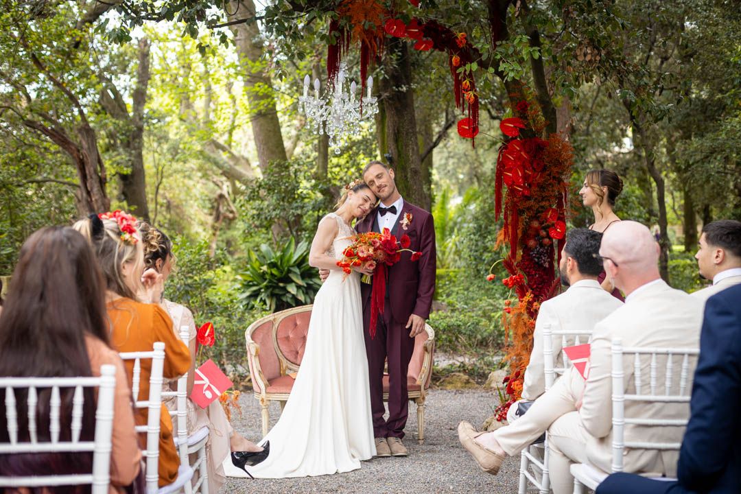 Outdoor garden wedding ceremony with red floral decorations and white chairs.