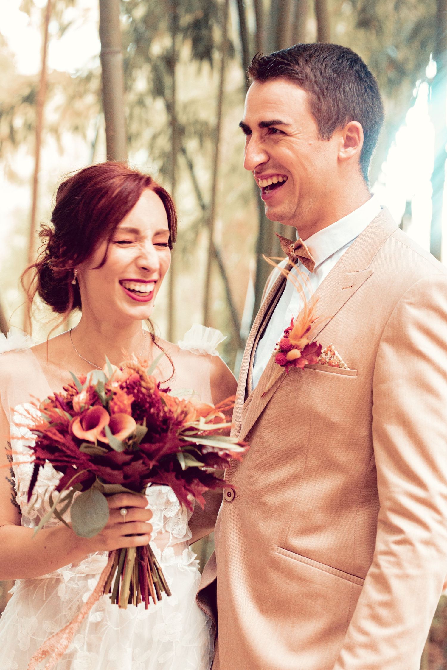 Wedding couple in light colors share joyful laughter while holding a burgundy bouquet outdoors.