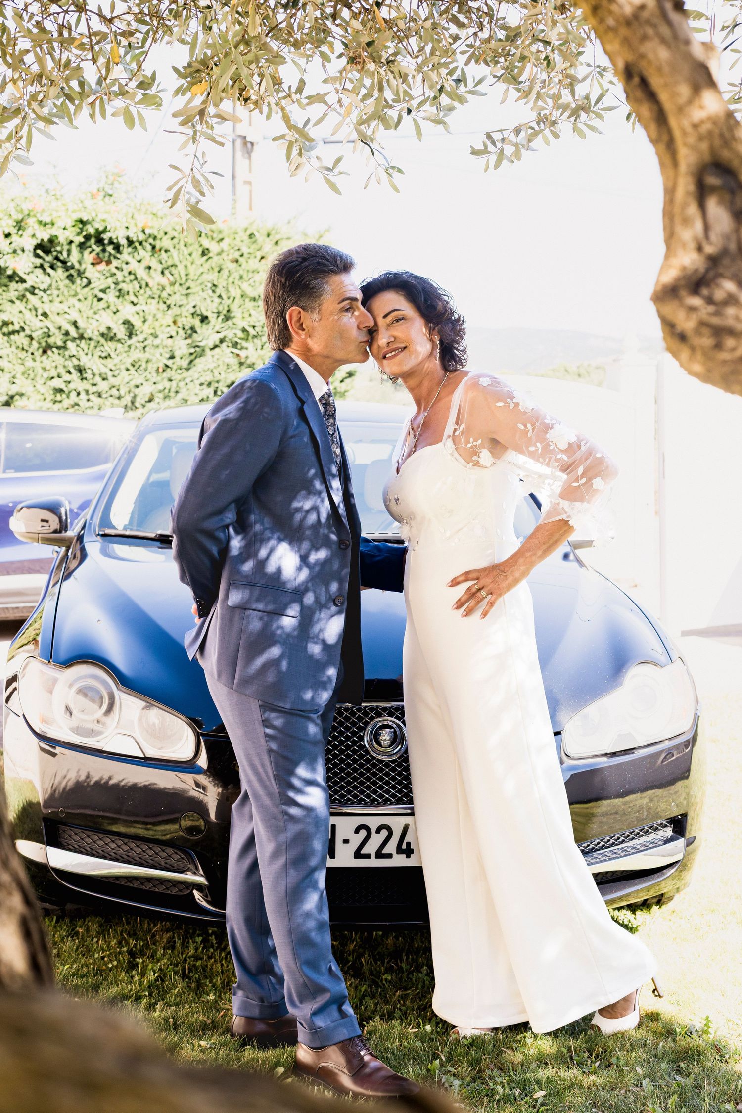 Elegant couple shares romantic moments near a luxury car in an outdoor garden setting.