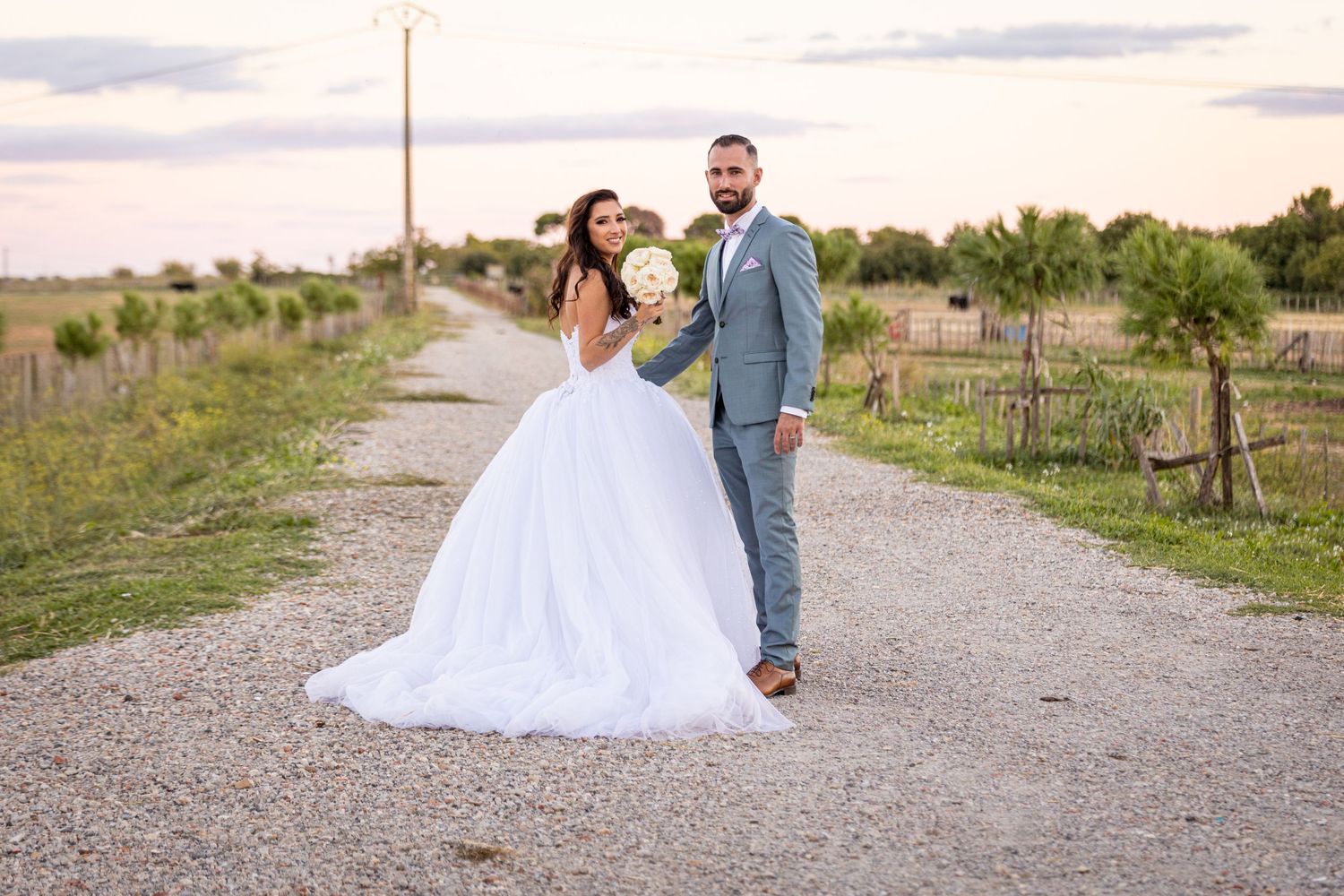 Wedding couple walking together on rustic path at dusk with string lights overhead.