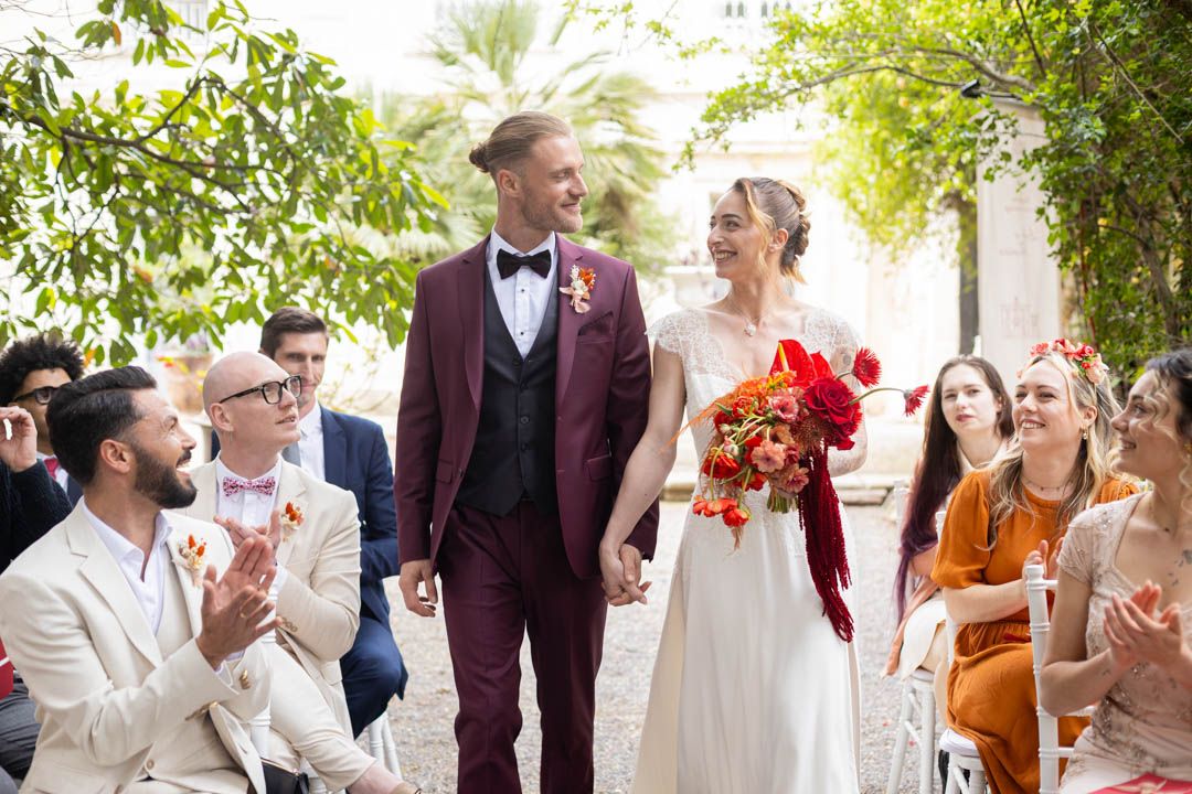 Wedding ceremony guests applaud as couple in burgundy suit and white dress walk down aisle.
