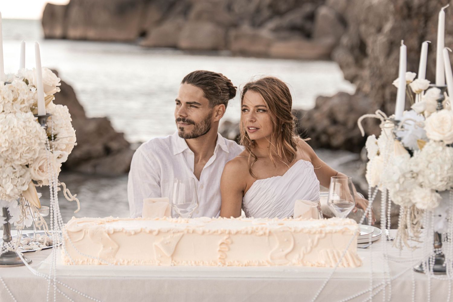 Elegant couple in white attire sitting at a decorated outdoor wedding table with a cake and floral arrangements by the sea.
