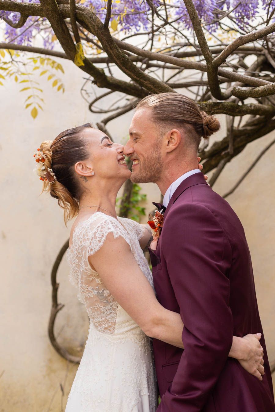 Intimate wedding moment captured under purple flowering wisteria branches in garden setting.