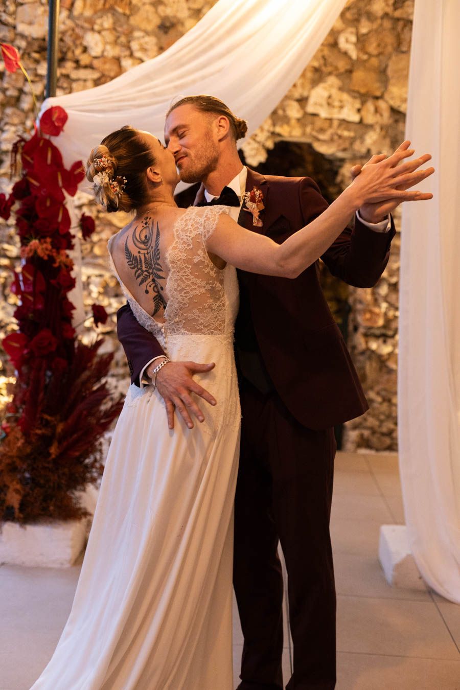 Couple dancing gracefully in front of stone wall with white draping and red floral arrangements.