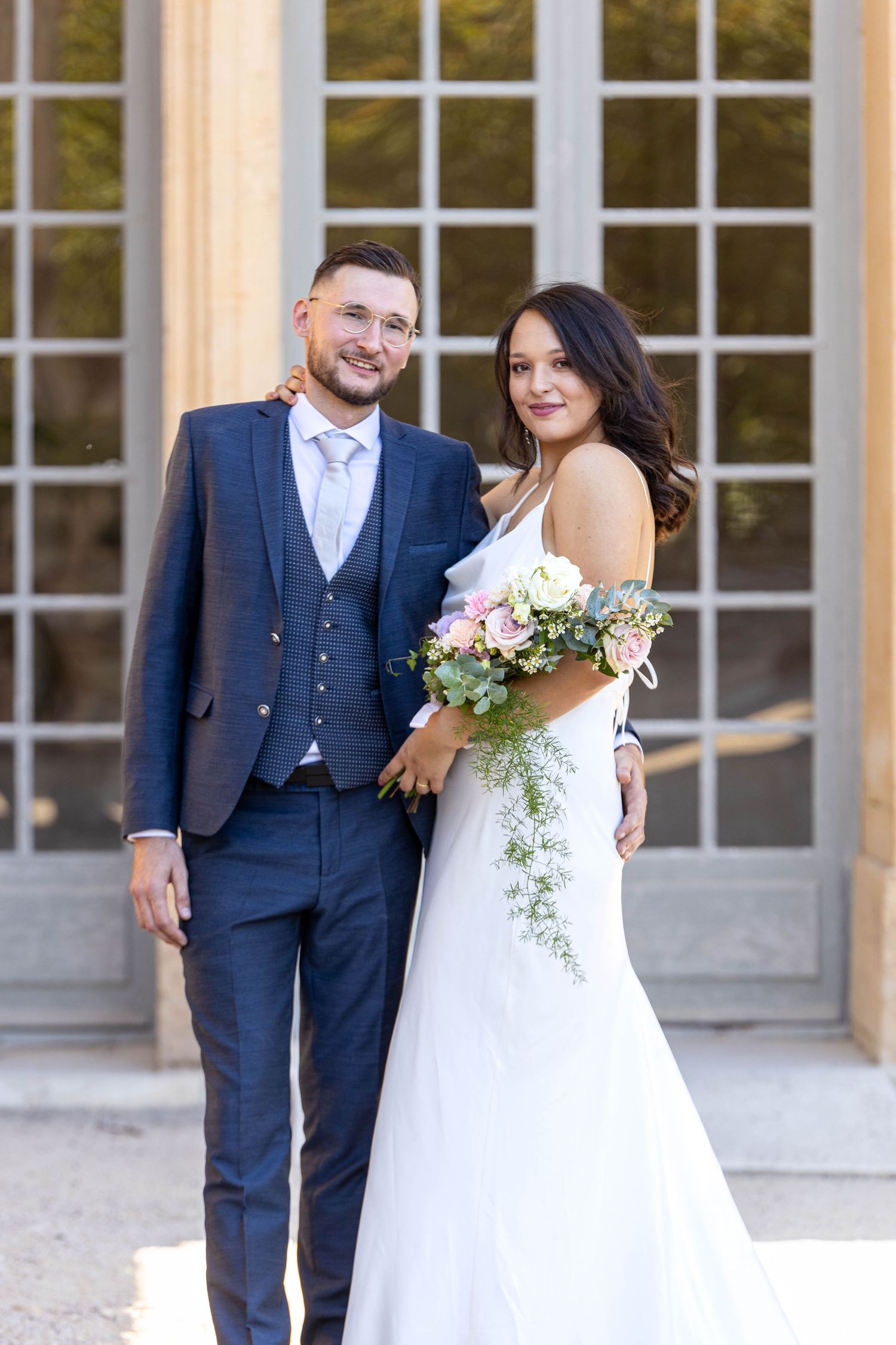 Elegant wedding couple poses in front of classic building with white paned windows.