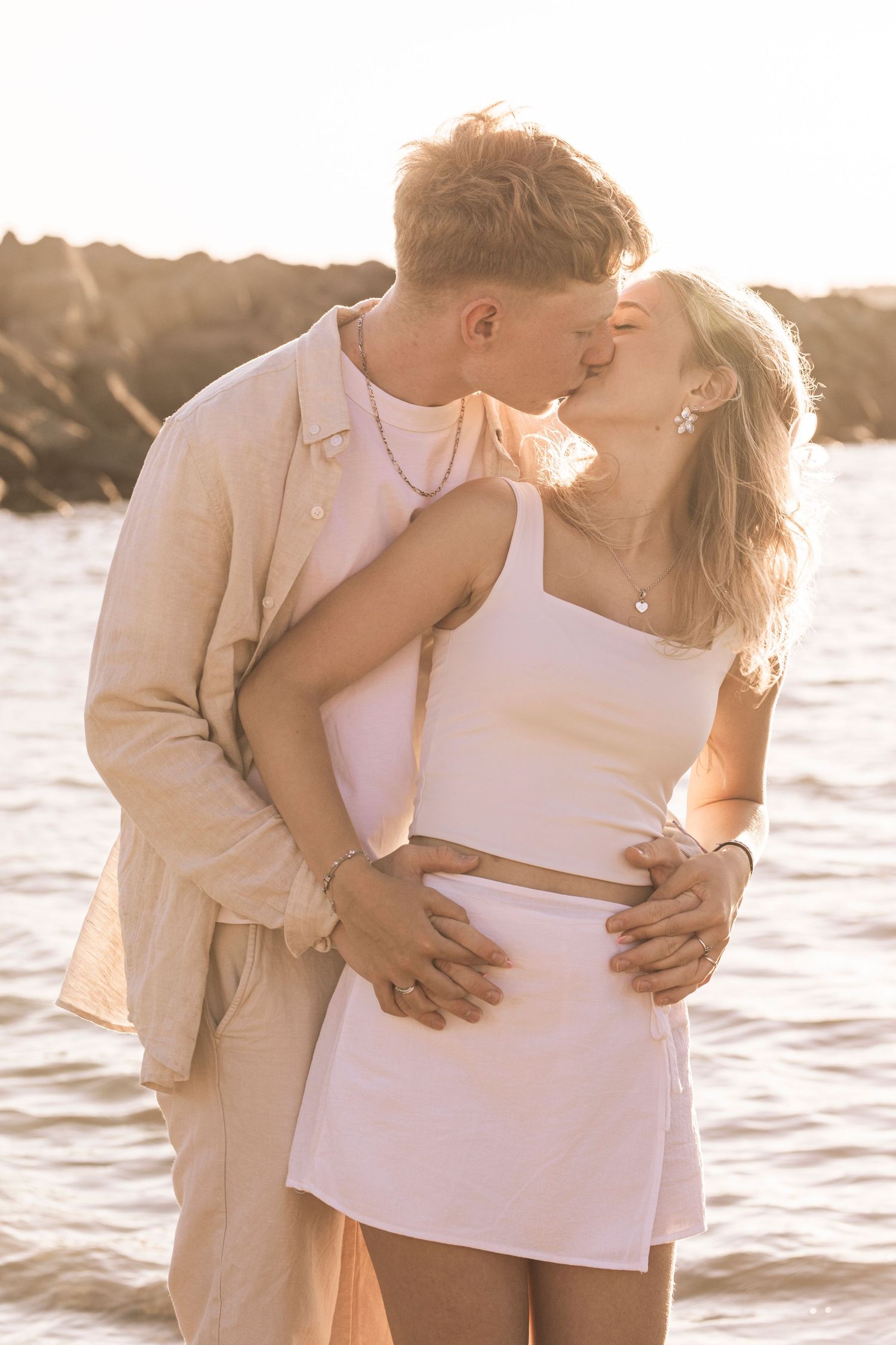 Romantic couple embraces on beach at golden hour with sunlit water in background, shot in warm sepia tones.