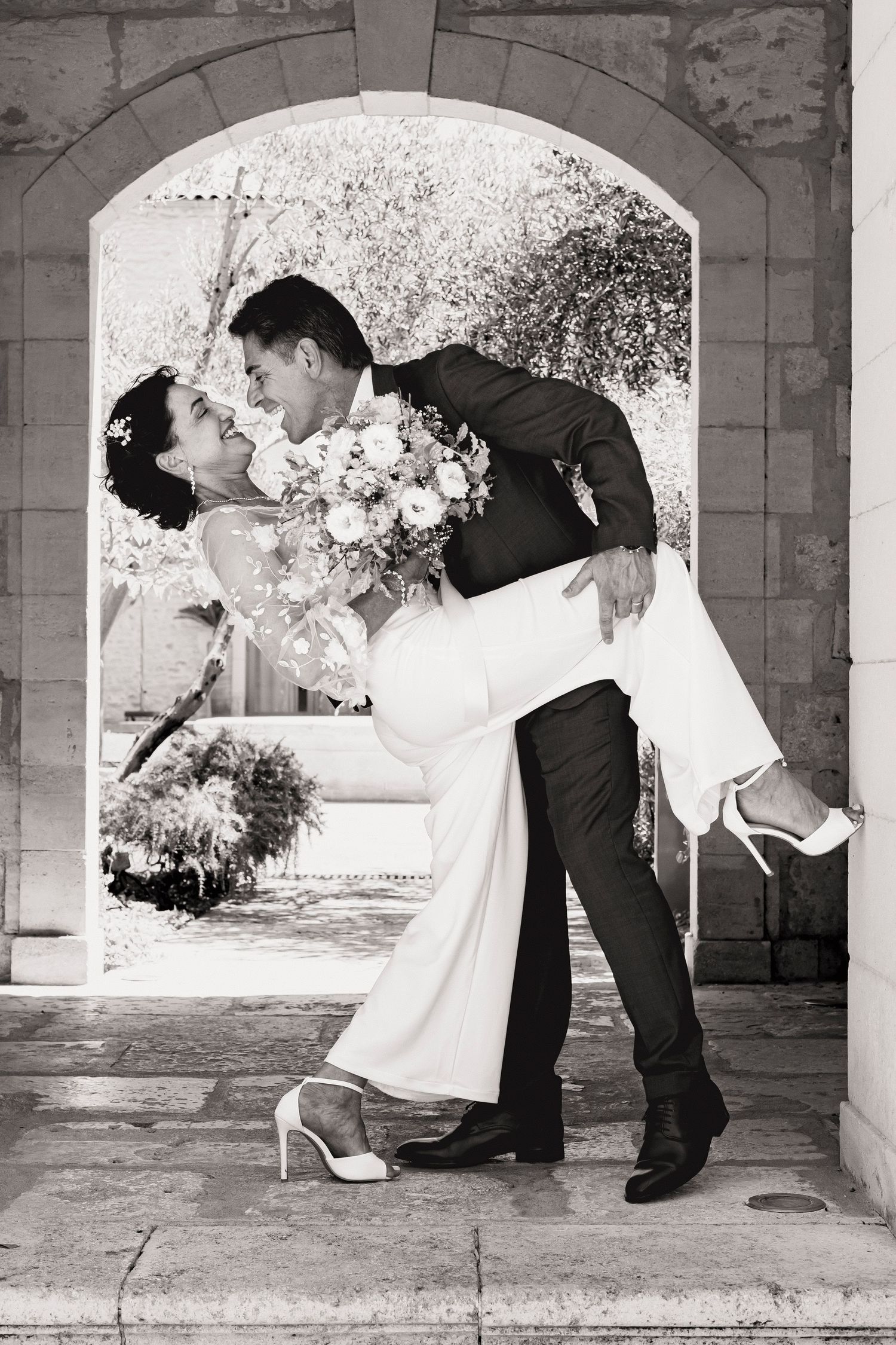 Black and white wedding photo shows dramatic dip pose under ornate stone archway.