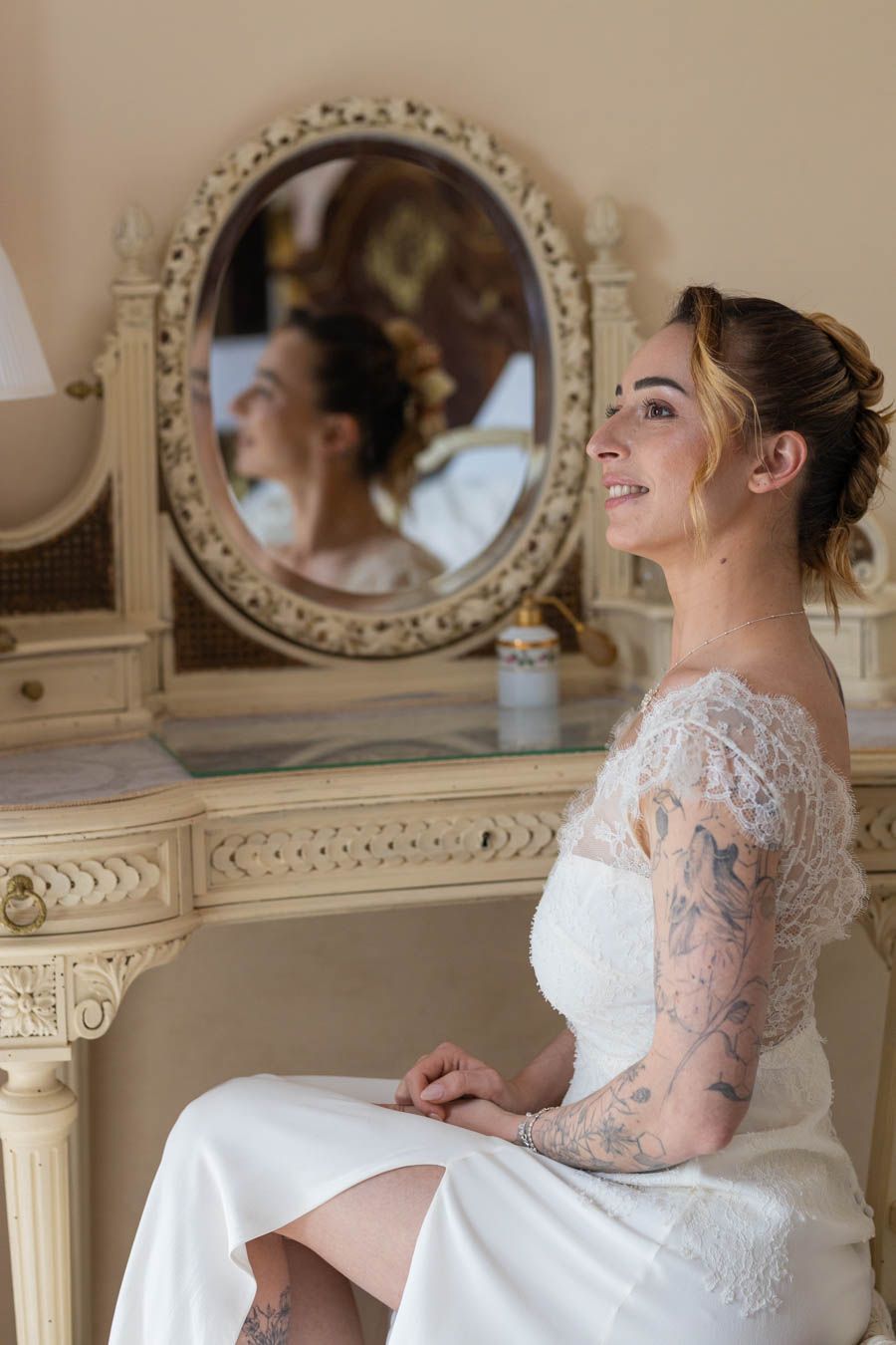 Elegant white lace wedding dress shown in a vintage-style dressing room with ornate mirrors.