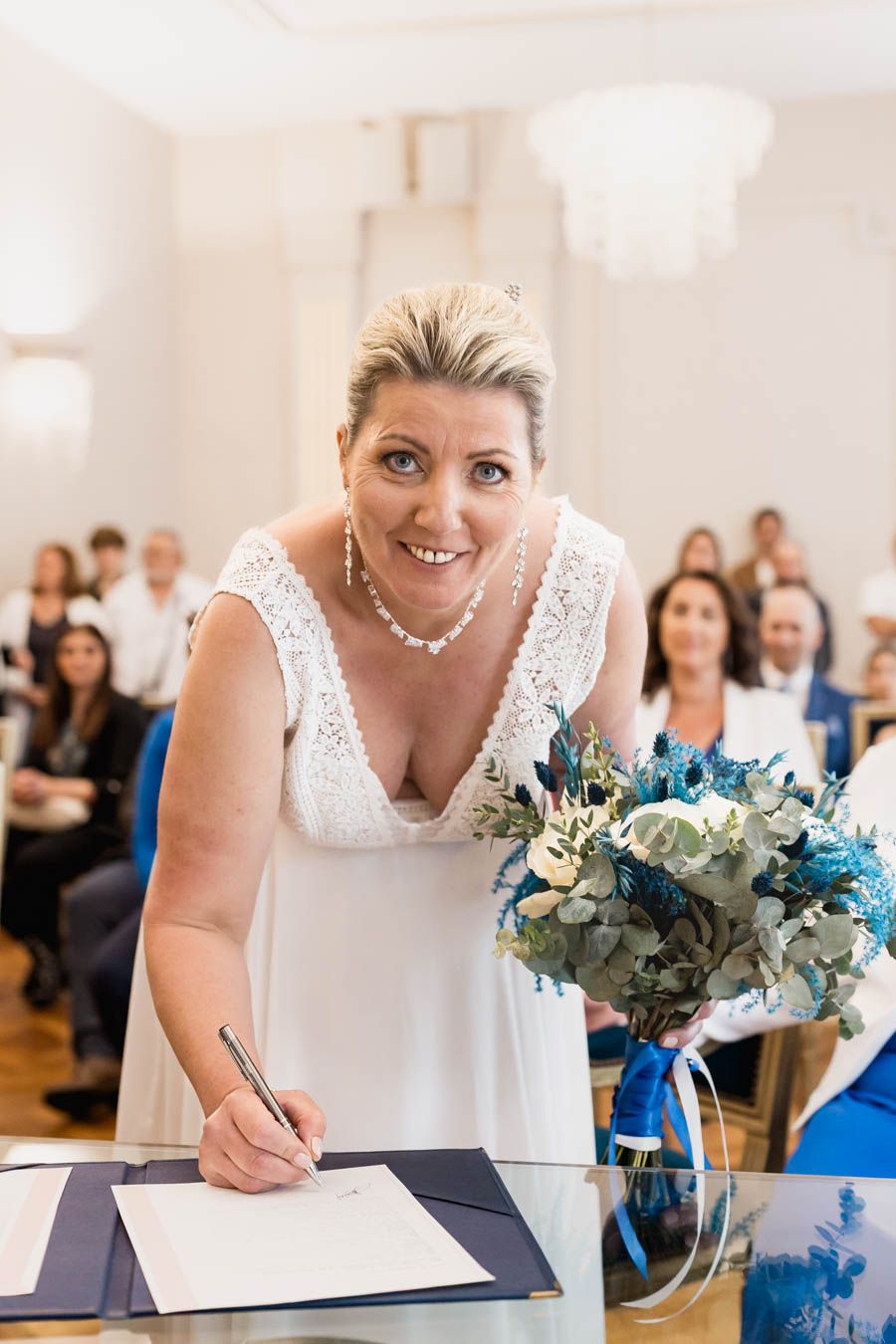 Bride in white dress signing marriage documents with blue bouquet on table.