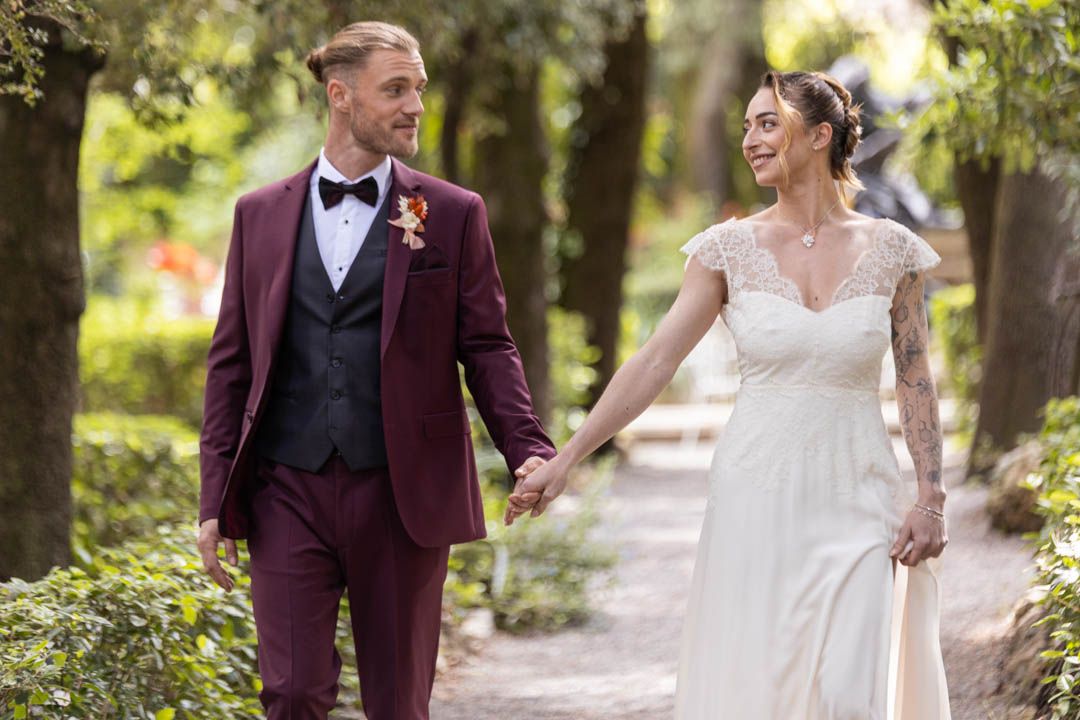 Couple walking hand in hand along a tree-lined path in wedding attire.