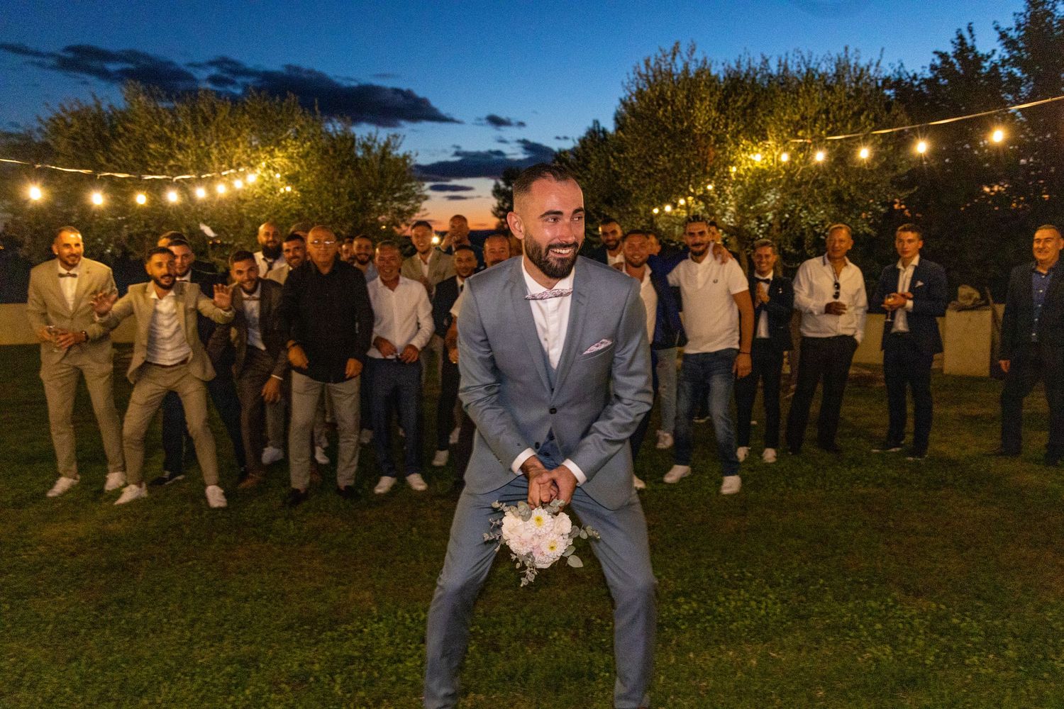 Outdoor wedding reception at dusk with string lights and group gathering on lawn.