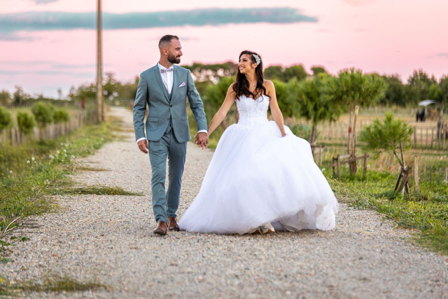 Wedding couple posing on gravel pathway during pink sunset with scenic rural backdrop.