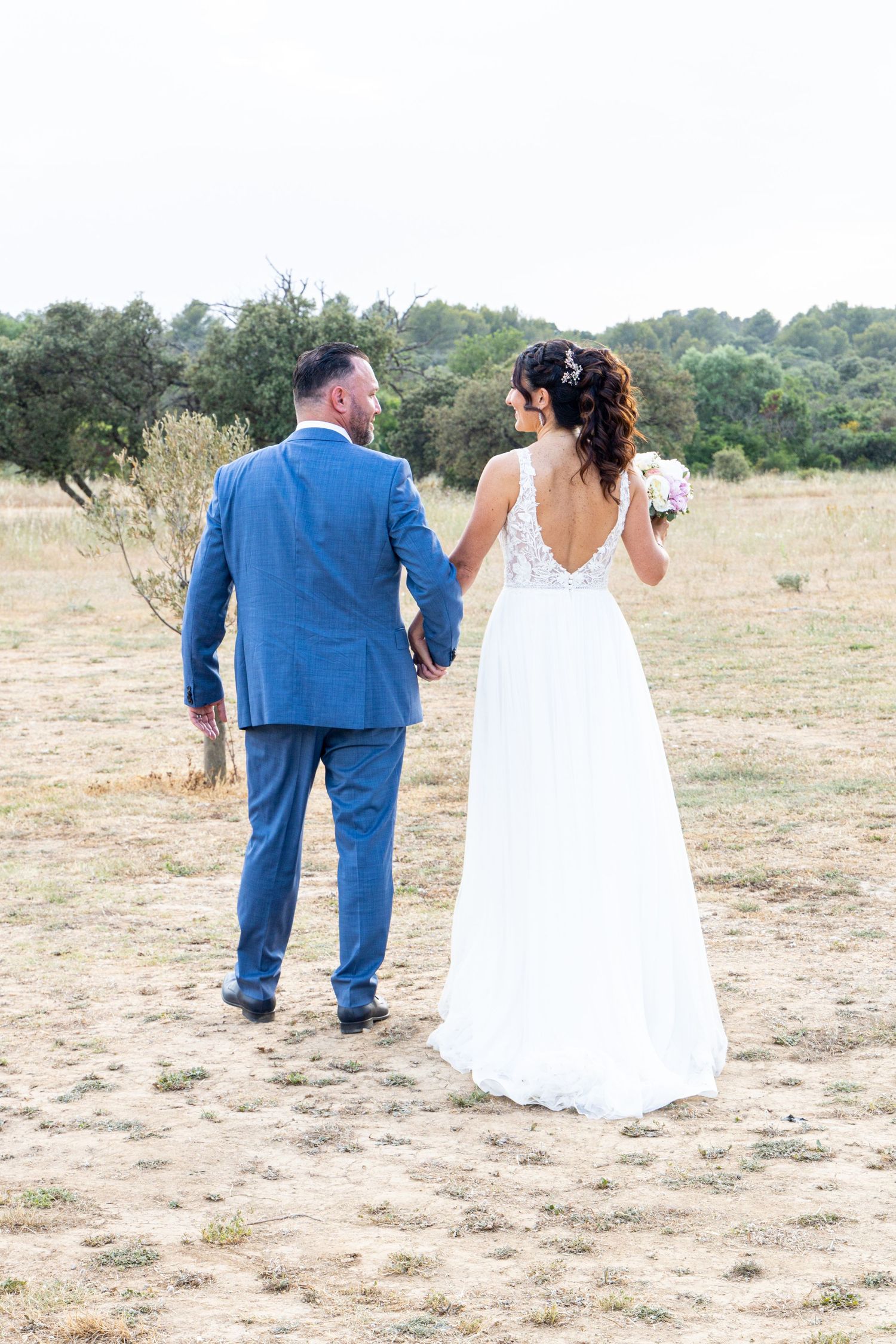 Walking couple in wedding attire against rustic outdoor backdrop.
