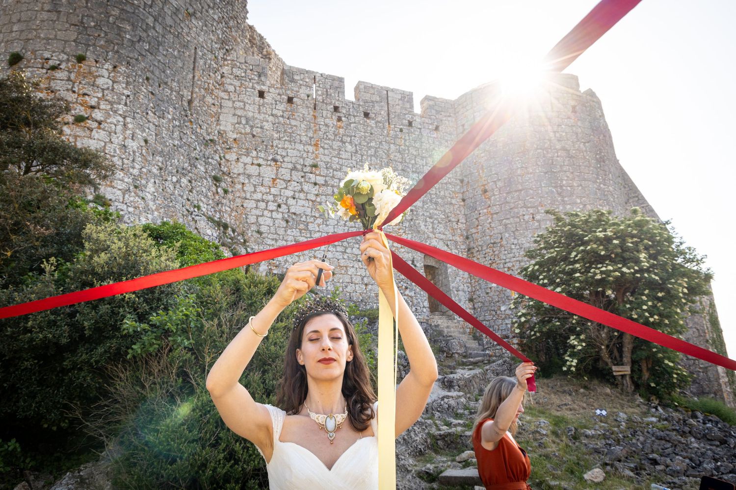 Bride reaching up to colorful ribbons against ancient castle walls during sunset.