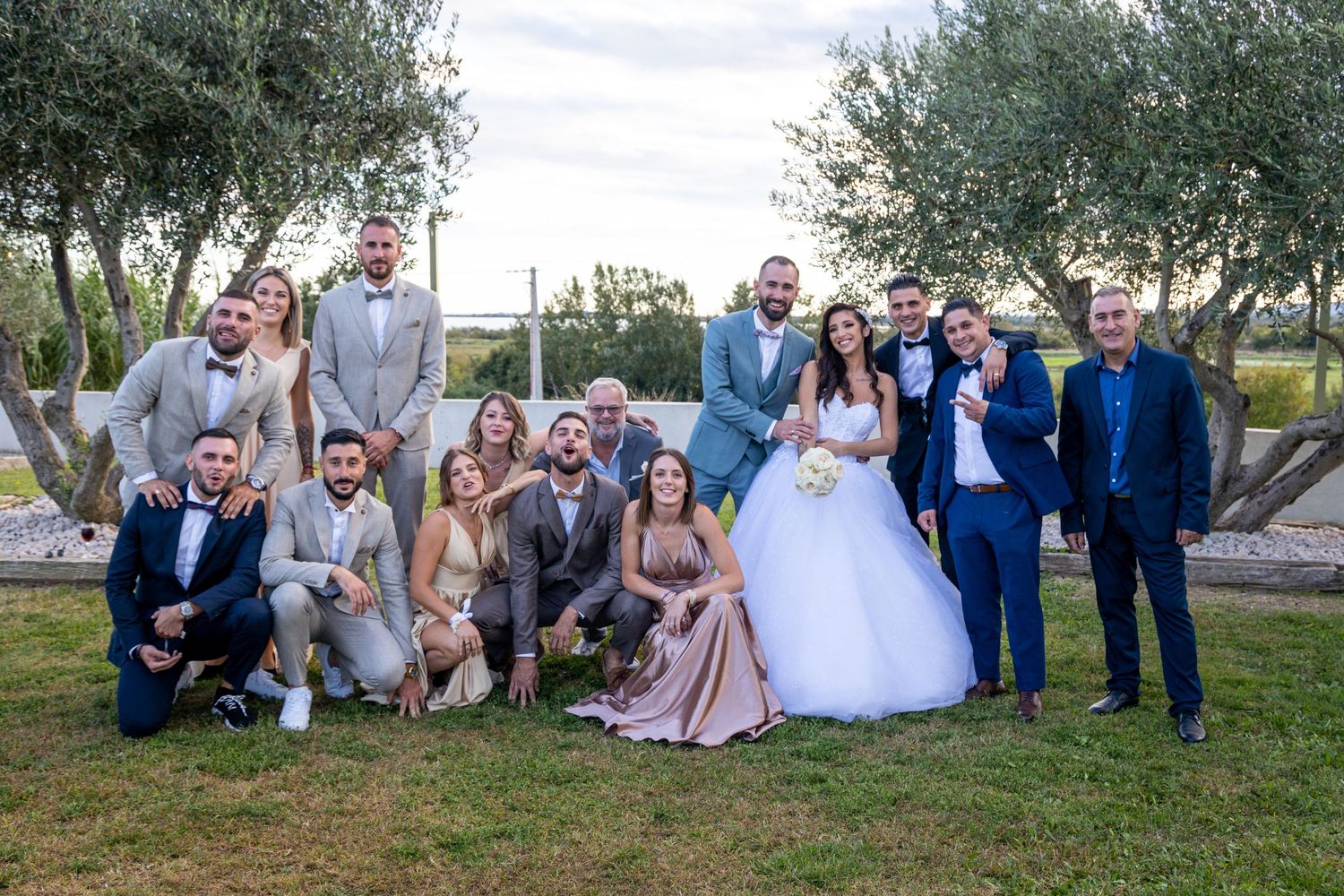 Wedding party posing for group photo outdoors with olive trees in background.