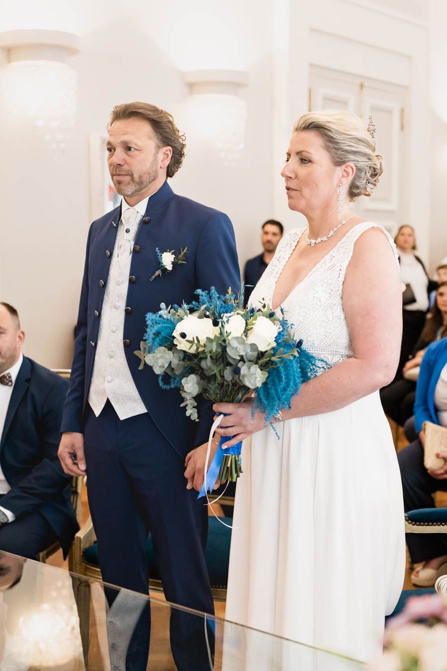 Wedding ceremony in bright indoor venue with couple and blue floral bouquet.