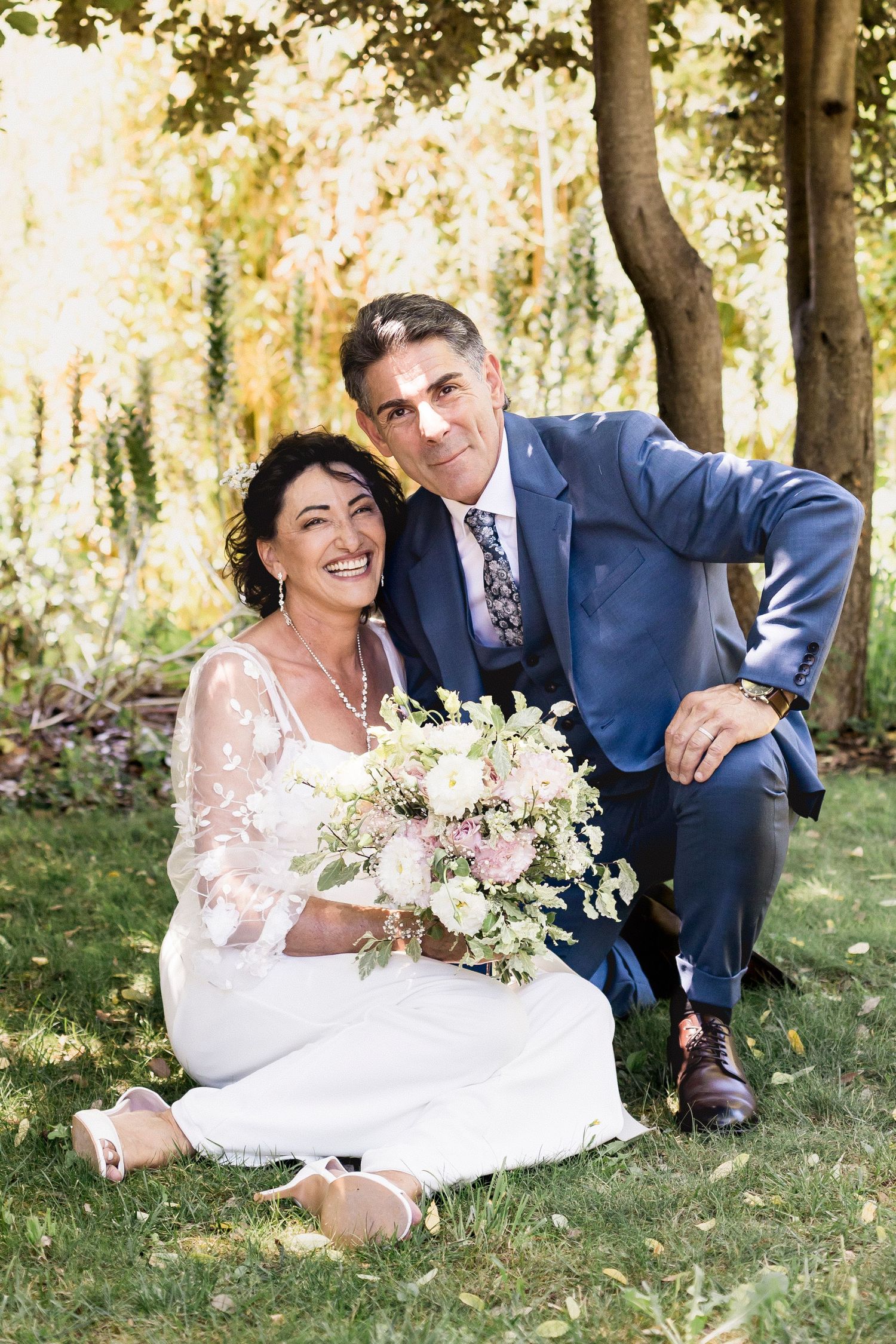 Joyful wedding portraits captured on grass under a canopy of trees.