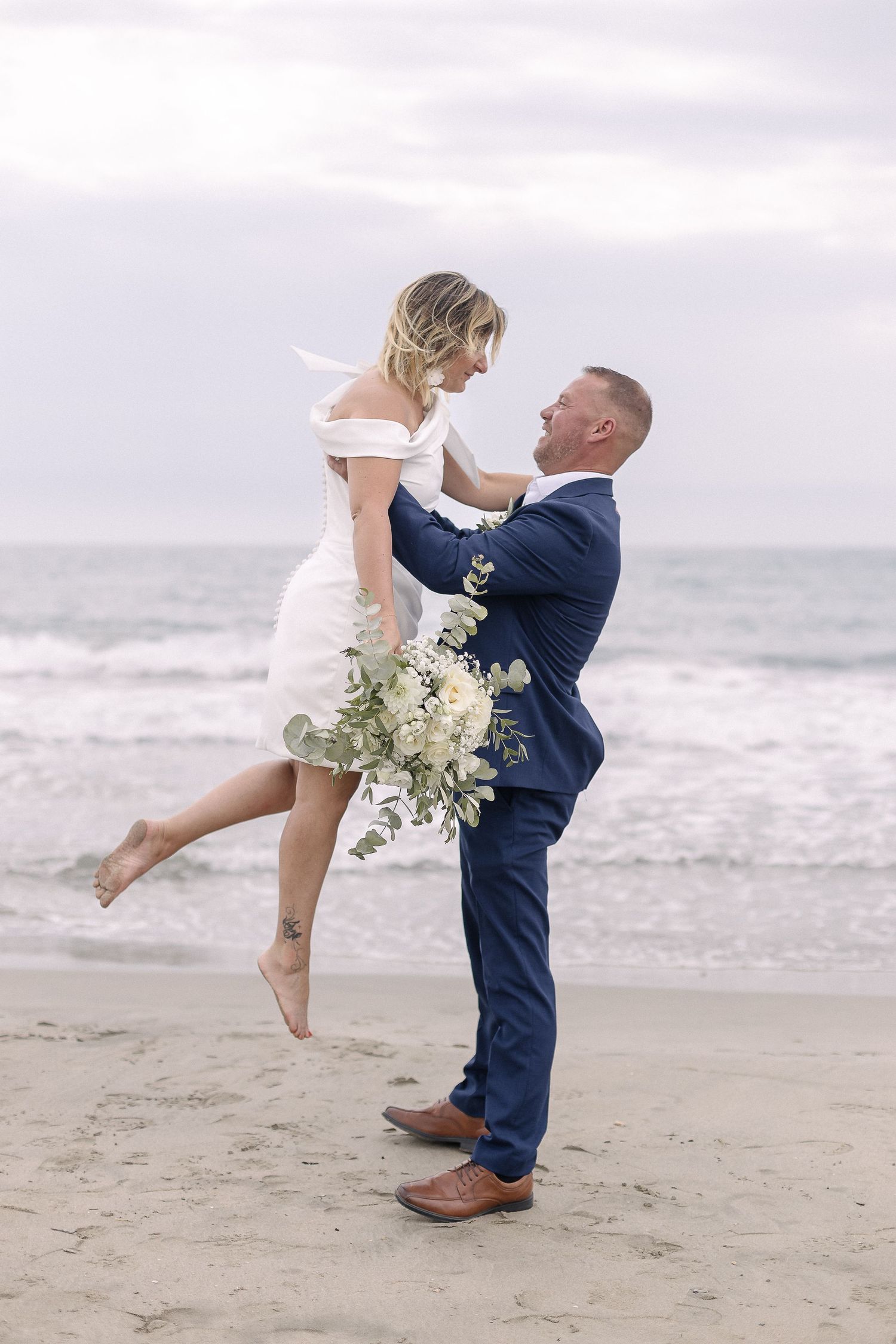 Playful beach wedding photo with couple dancing in sand, bride in white, groom in navy suit.