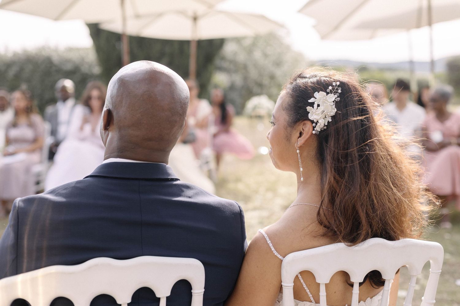Wedding couple seated in white chairs during an outdoor ceremony.